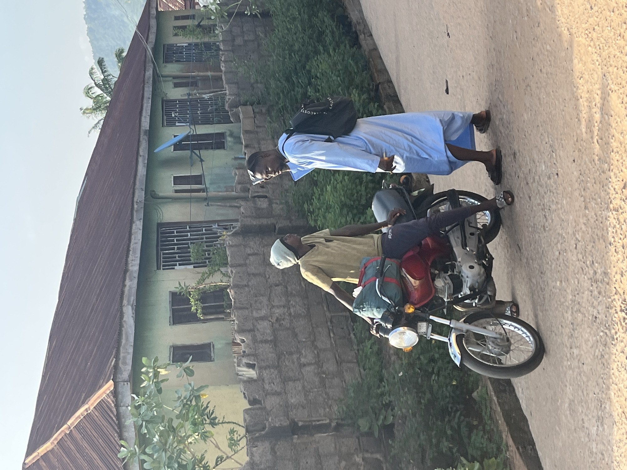In a sunlit outdoor scene, two young men are present in a residential area. The man on the left is seated on a red motorcycle, wearing a light brown t-shirt and a light-colored headscarf, with a green duffel bag strapped to the motorcycle's rear. The man on the right stands, dressed in a light blue robe and wearing a black backpack, and appears to be speaking on a mobile phone. Behind them, a low, rough stone wall separates the street from a pale green, single-story building with a corrugated metal roof. The building has barred windows and a satellite dish attached. Lush green vegetation grows between the wall and the building, and also along the side of the road. The ground is a paved, sandy surface. The sky is clear and bright.