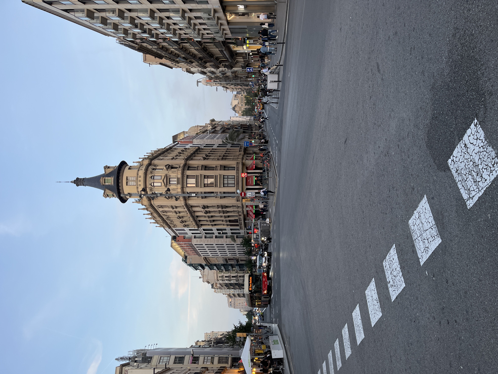A vertically oriented photograph captures a bustling city street in Barcelona, Spain, stretching from the foreground towards the distance. On the left side of the street, a grand, ornate building with a distinctive dark, pointed dome and turret stands out, its facade adorned with numerous windows and balconies. Adjacent and beyond it, various other multi-story buildings line the street, showcasing a mix of architectural styles. On the right side of the street, another continuous row of buildings features ground-level storefronts and residential units above.
The street is populated with people and vehicles, indicating an active urban environment. Pedestrians are visible on both sidewalks, walking, standing, or gathering. On the right sidewalk, an outdoor dining area under a white tent provides seating for patrons. A red double-decker bus, marked with "555 Pere Maragall" (or similar, like "Port Forum") on its destination sign, is prominent on the right lane of the street, heading away from the viewer. Other vehicles, including cars and parked motorcycles and bicycles, are also present.
The clear, light blue sky with a gentle, warm glow suggests the time of day is late afternoon or early evening, with artificial lights starting to become visible on buildings and street signs. The asphalt road features white dashed lines and distinctive cracked white square markings on its right side. Notable visible text includes the "Sabadell" bank logo on a storefront and "loko" on a sign near the outdoor cafe. The architectural style, the specific bus line, and the visible bank logos strongly confirm the location as Barcelona.