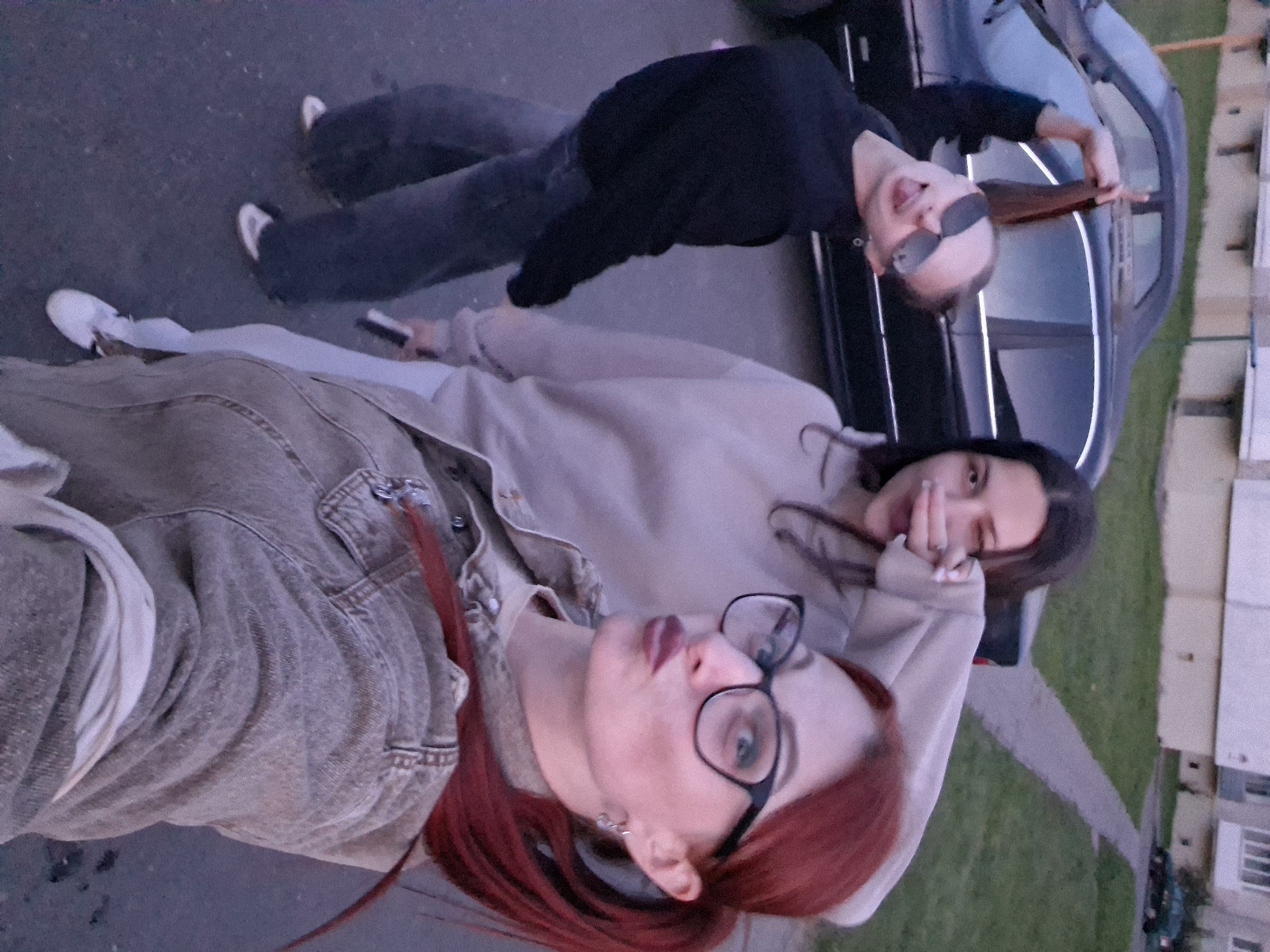 Three women are in an outdoor setting, appearing to be in a residential area of Myadzyel, Belarus. The woman in the foreground, with red hair and glasses, is taking a selfie, puckering her lips. She is wearing a denim jacket over a light-colored garment. To her left, a woman with dark hair is wearing a light-colored hoodie and has her hand near her face. To the far left, a third woman with blonde hair, wearing sunglasses and a dark sweatshirt, is sticking out her tongue and holding her hair up. She is wearing jeans and white sneakers. A black car is parked behind the women, and a building with windows is visible in the background, along with a paved walkway and a grassy area. The lighting suggests it is late afternoon or early evening.