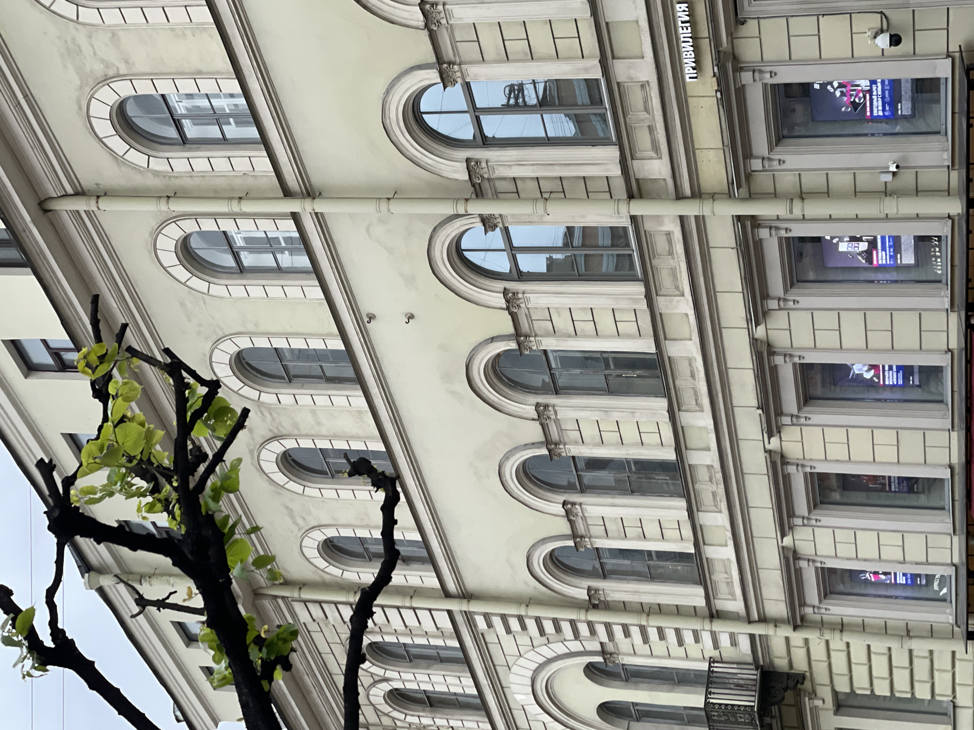 The image shows the exterior of a building in Unknown, Unknown. The building features a light-colored facade with multiple rows of arched windows. A tree with green leaves is partially visible in the foreground, obscuring the lower left corner of the building. A drainage pipe runs vertically along the facade. There are also some rectangular windows with posters visible on the right side of the building. A security camera is mounted near the upper right corner. The building's name "ПРИВИЛЕГИЯ" is seen on the top.