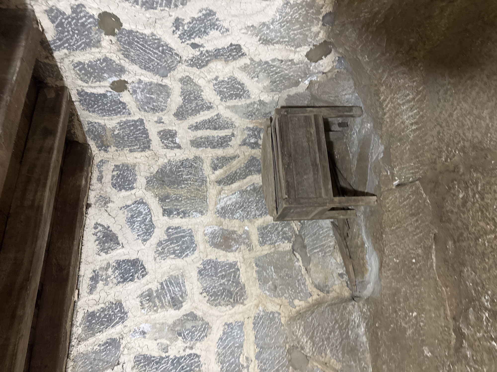The scene depicts the interior of a structure, likely a building or cave, in the Sà Phìn Commune of Vietnam. The walls are constructed of rough-hewn stones, arranged in an irregular pattern. A small, wooden box is affixed to one of the walls. A wooden beam is partially visible. The environment appears dark and possibly damp.