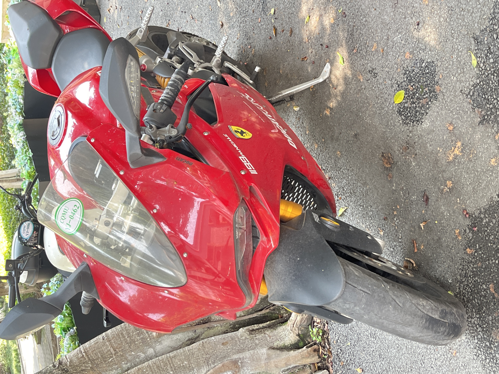 A bright red Ducati 1199 Panigale motorcycle is parked outdoors, angled diagonally with its front wheel pointing towards the right side of the frame. The motorcycle's kickstand is extended, supporting the bike. The setting appears to be a paved area, likely asphalt, scattered with dry leaves. In the background on the left, green and light blue foliage is visible, suggesting a natural or landscaped area. The scene is illuminated by natural daylight, indicating it is daytime.
Notable details include the clear branding on the motorcycle: "Ducati" is visible near the engine, "1199 PANIGALE" is prominently displayed on the side fairing, accompanied by a yellow shield logo. Two green circular stickers are affixed to the windscreen, one clearly readable as "COMU YJ-0462" and another partially visible as "COMU YJ-0080". No people are visible in the image, and there is no discernible activity other than the motorcycle being parked. The location is Yuzhong District, China.