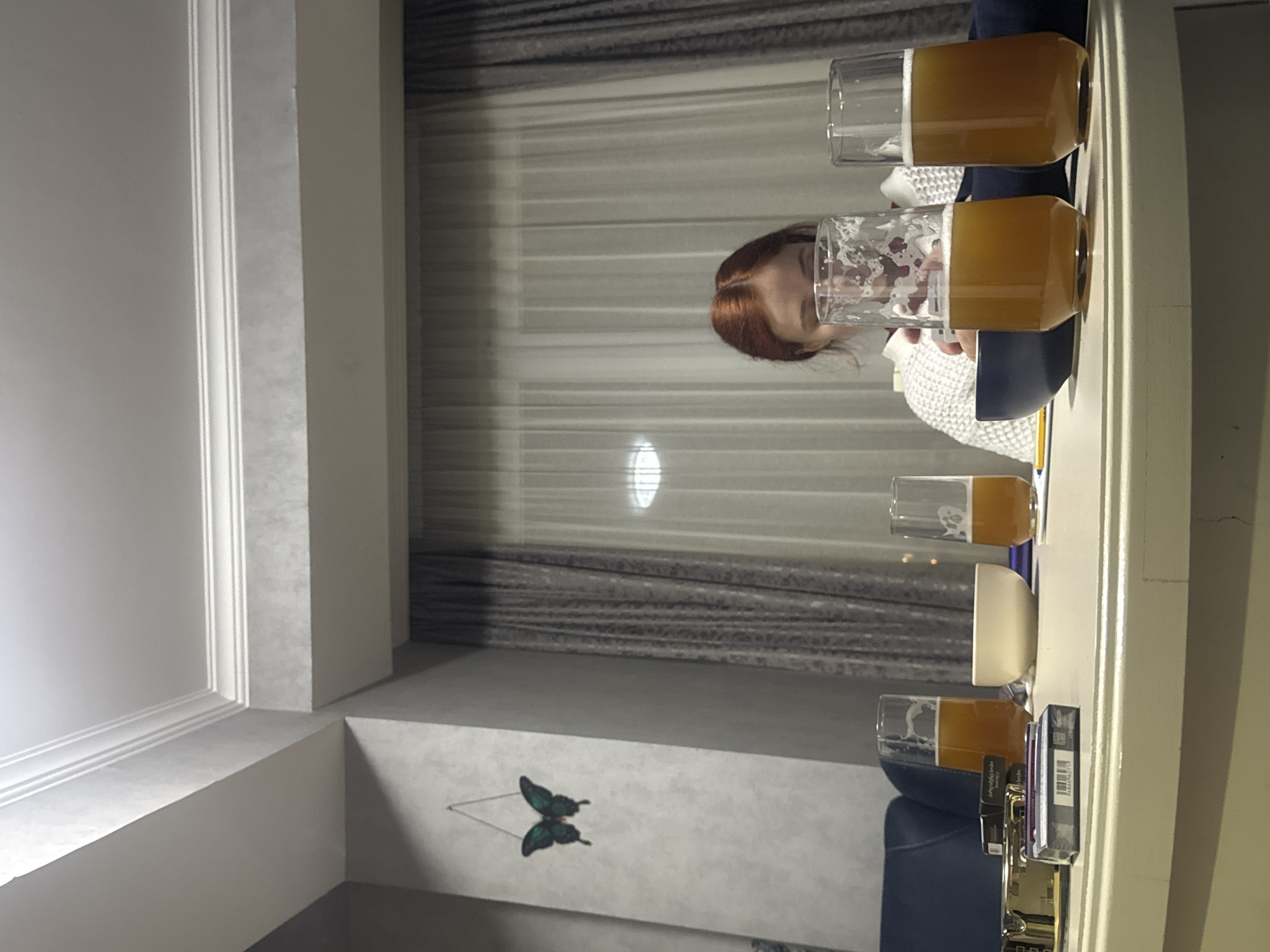 The photograph captures a woman sitting at a window in Tbilisi, Georgia, engaged in what appears to be a beer-tasting session. Two tall, filled glasses of beer are positioned on the windowsill, while a third glass sits slightly further away. The woman, with red hair, is facing the window, her face partially obscured by one of the beer glasses. She is wearing a white sweater and a dark blue jacket. The window overlooks a building interior. A decorative butterfly is positioned near the bottom of the window and a lamp is sitting on the windowsill. Additional items, including what appear to be cards or packaging materials, are also present on the windowsill. The room has a neutral color scheme, with white walls and grey curtains.