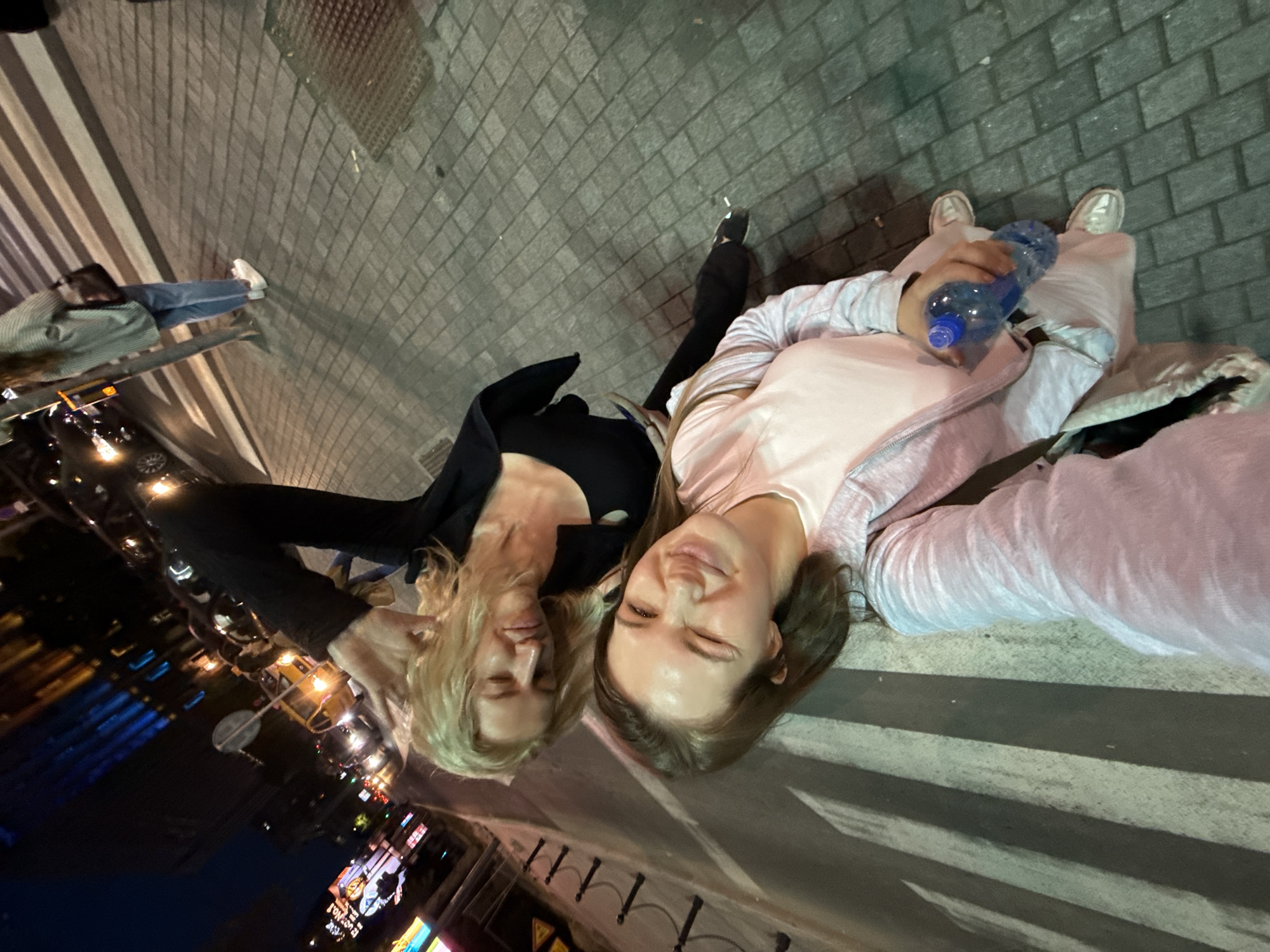 Two women are lying on a crosswalk at night. The crosswalk is in Belgrade, Serbia, indicated by the street lights and buildings in the background. The woman on the left has blonde hair and is wearing a black top. The woman on the right has brown hair and is wearing a pink shirt with a gray jacket. She is holding a plastic water bottle. One person is seen walking in the background, and cars are also visible. The ground is paved with gray bricks.