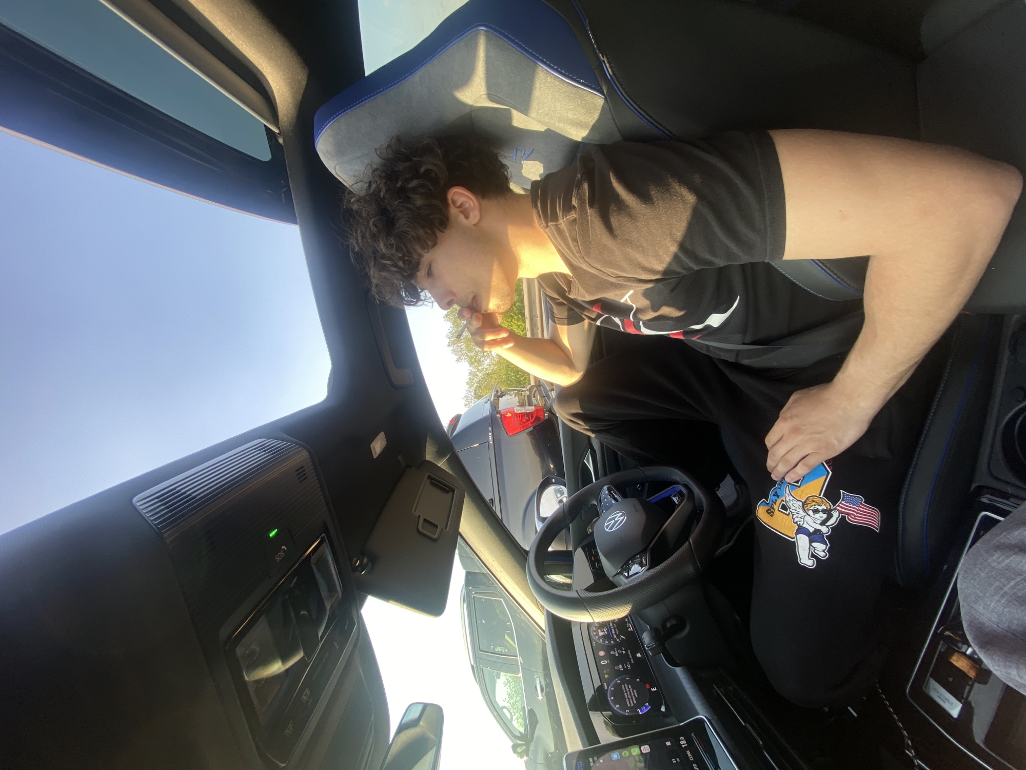 The image shows a young man seated in the driver's side of a modern car, bathed in bright sunlight. He has curly brown hair and is wearing a black t-shirt with a partially visible graphic and black sweatpants. He is holding a white object, possibly a cigarette or vape pen, to his mouth.
The car's interior is visible, featuring a sporty seat with blue and gray upholstery. The dashboard includes digital displays, with one showing "18.0 C/Q", likely indicating temperature. A multi-function steering wheel, possibly with a VW logo, is in front of him. Above, an overhead console includes ventilation grilles and an "SOS" button illuminated with a green light. A panoramic sunroof is visible overhead, and a sun visor is lowered on the driver's side.
The man's black sweatpants prominently display a large graphic on the left thigh: an angel figure wearing sunglasses and holding an American flag, set against a yellow and blue design that resembles the letters "LA" or "UA."
Outside the car, under a clear blue sky, parts of another dark-colored vehicle and some trees are visible through the windows. The strong sunlight and clear sky indicate daytime. The scene suggests a relaxed moment, possibly while parked in Wroughton, United Kingdom.