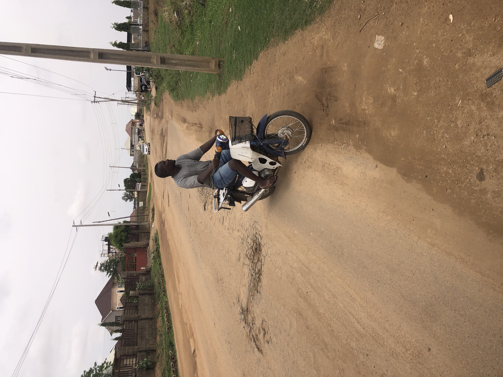 A man rides a small motorbike on a dirt road in a residential area. The road is dusty with several potholes. There are power lines and wooden utility poles along the side of the road. Houses with fenced yards are visible in the background. The scene is in Hanwa, Nigeria.