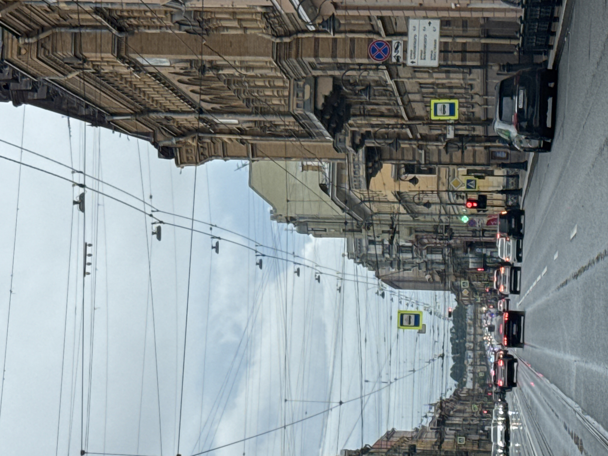 The image shows a wide street in Saint Petersburg, Russia, likely Nevsky Prospekt, given the architecture and overhead wires. The street is lined with ornate, multi-story brick buildings, featuring decorative window frames and balconies. Numerous overhead wires, likely for trolleybuses or trams, crisscross the sky, creating a dense network against a cloudy sky. Traffic is moderate, with several cars visible on the wet asphalt road, suggesting recent rain. Traffic lights, some red and some green, are present, as are street signs and pedestrian crossing indicators. The overall atmosphere is urban and overcast, characteristic of Saint Petersburg's climate.