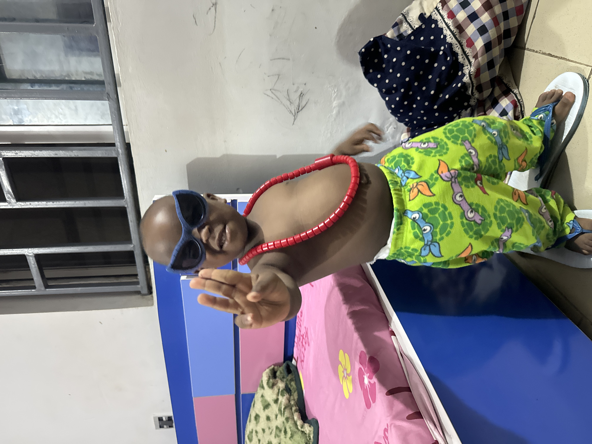 A young child is lying on a pink bed in an indoor environment. The child is wearing blue sunglasses, a red beaded necklace, and green pajamas patterned with cartoons. They are also wearing white and blue flip-flops. The bed is next to a wall with a window featuring horizontal metal bars. There's also a patterned fabric or pillow near the bed. The location is Sabon Tasha, Nigeria.