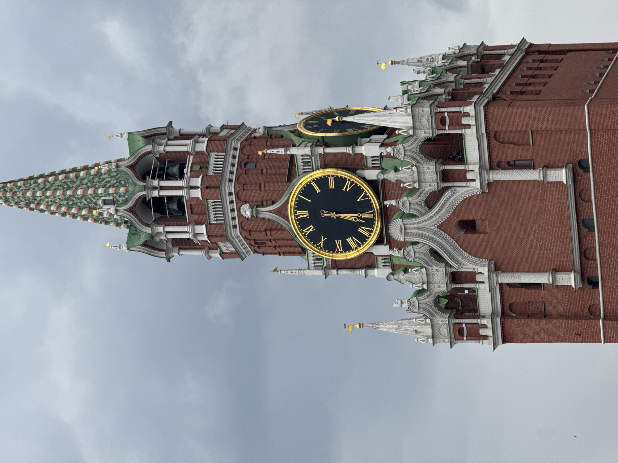 The image shows a close-up, low-angle, outdoor, eye-level shot of the Spasskaya Tower of the Moscow Kremlin, Russia, against a cloudy, overcast sky. The iconic red brick tower features intricate white detailing, green roofs, and a decorative spire with colorful tiles. Prominently displayed on the tower are two large, round clocks with Roman numerals and gold accents. The clock on the left shows the time as approximately 1:55, with the hour hand near the 2 and the minute hand nearing the 12. The clock on the right is partially obscured but appears to show a similar time. Several bells are visible through arched openings in the tower. The architecture is elaborate, with numerous spires, decorative elements, and arches. There are no people visible in the immediate frame of the tower. A tiny bird is visible in flight in the lower left portion of the image.