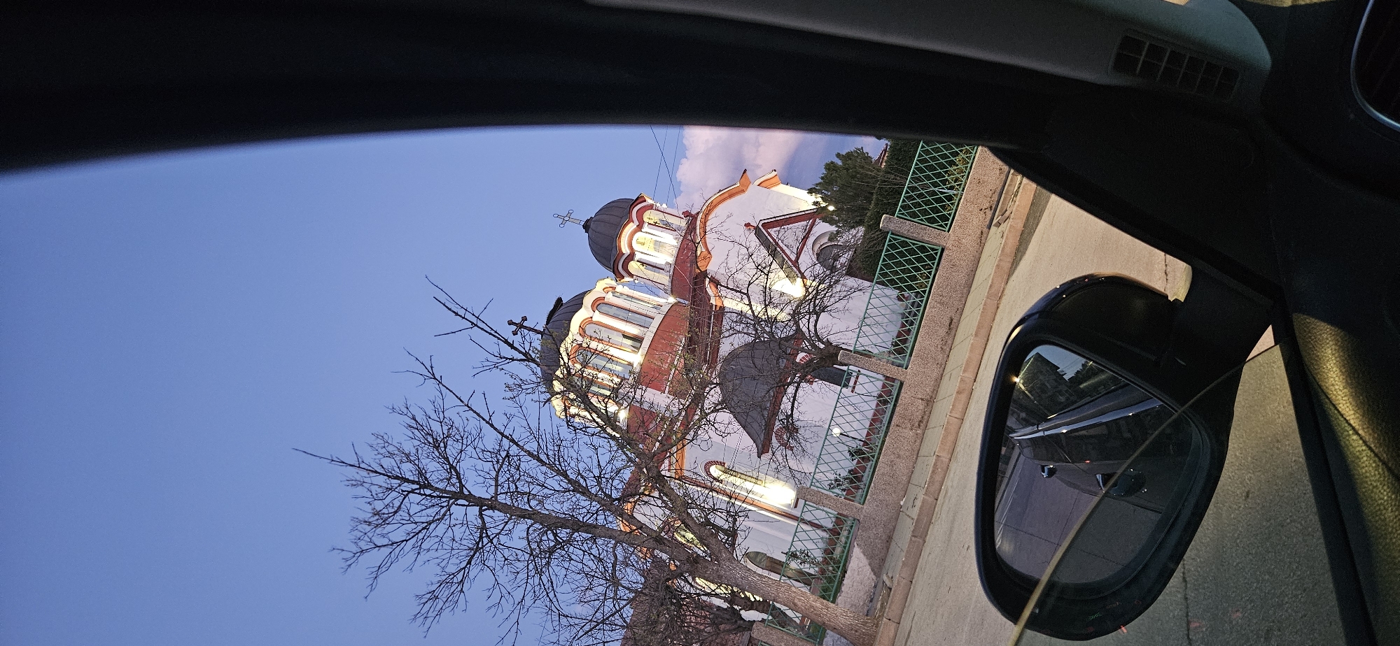 The image, taken from inside a car, shows the Church of the Holy Mother of God in Kostenets, Bulgaria. The church, a white and red building with dark gray domes topped with crosses, is partially obscured by a bare tree in the foreground. A green fence surrounds the church grounds. The sky is a clear blue, indicating twilight. The car's side mirror is visible in the bottom right, reflecting the road and nearby buildings. The interior of the car is dark, with the dashboard and window frame visible.