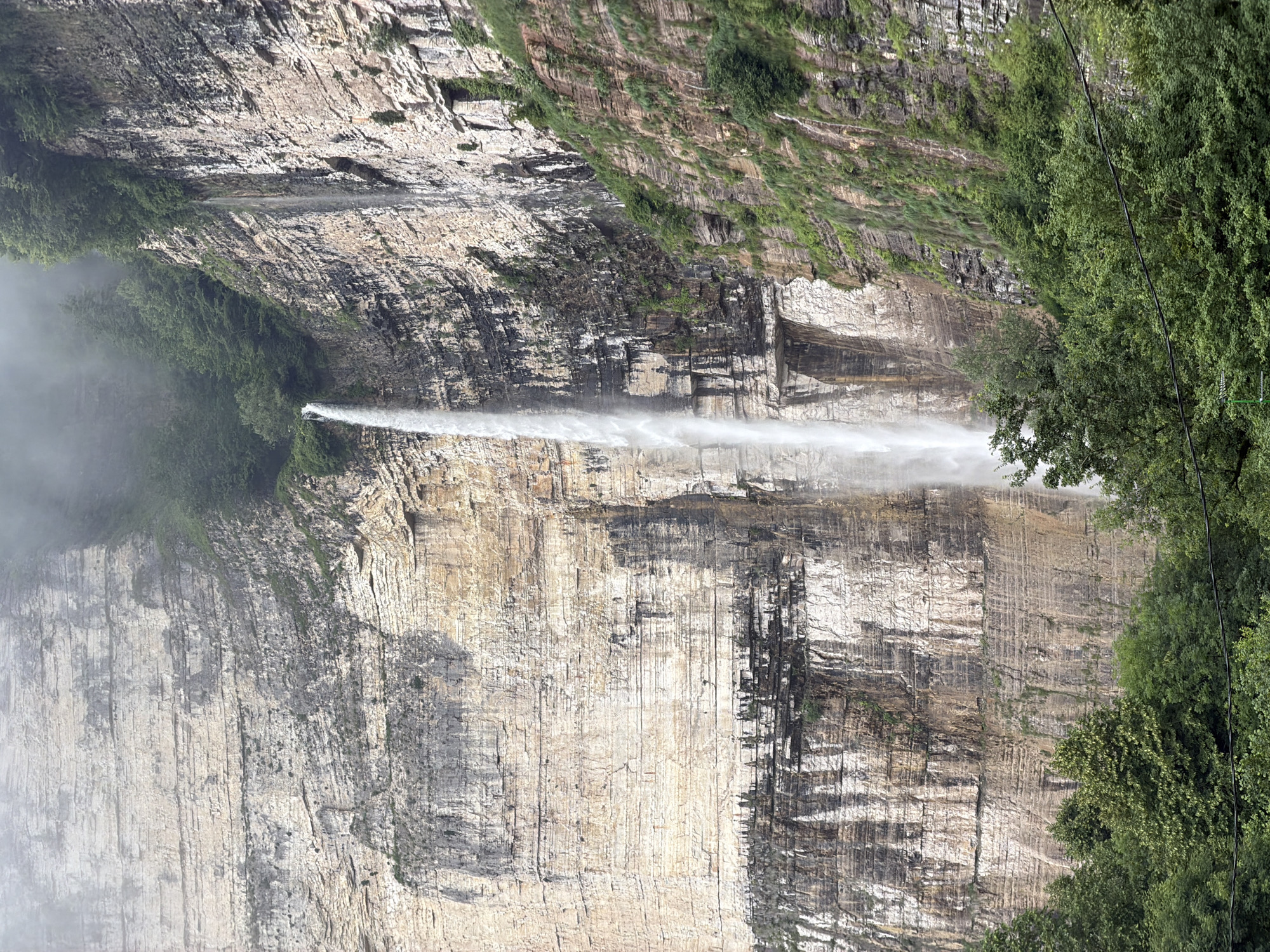 The image depicts a tall waterfall cascading down a sheer, layered rock cliff face. The cliff is a mix of light and dark rock striations, with patches of dark staining and greenery clinging to its surface. Trees and dense foliage are visible at the base of the cliff and on its upper edges, partially obscured by mist or fog. The waterfall itself is a thin, white stream of water, appearing to originate from the misty top of the cliff and plunging into the unseen area below. This is the Kinchkhaperdi Waterfall, located in Kinchkhaperdi, Georgia. There are no people or animals visible in the image. The overall environment suggests a natural, rugged landscape with a dramatic geological feature.