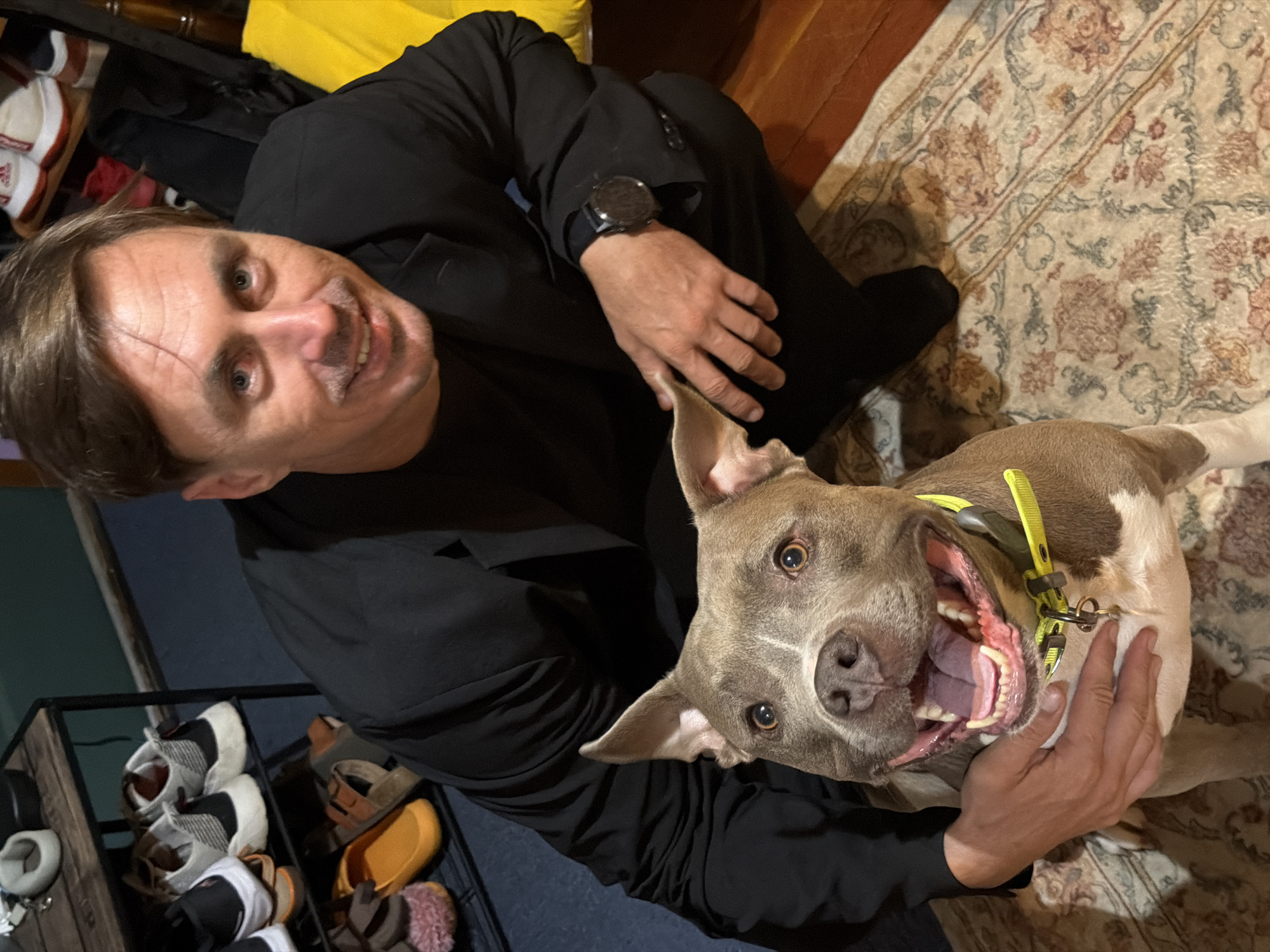 A man and a dog are posing for a picture in what appears to be a living room. The man is wearing a black suit and the dog is a light brown pit bull. The man is kneeling and petting the dog. The background is a rug with a floral pattern. There is a shoe rack in the background with several pairs of shoes on it. The image is taken from a slightly high angle.