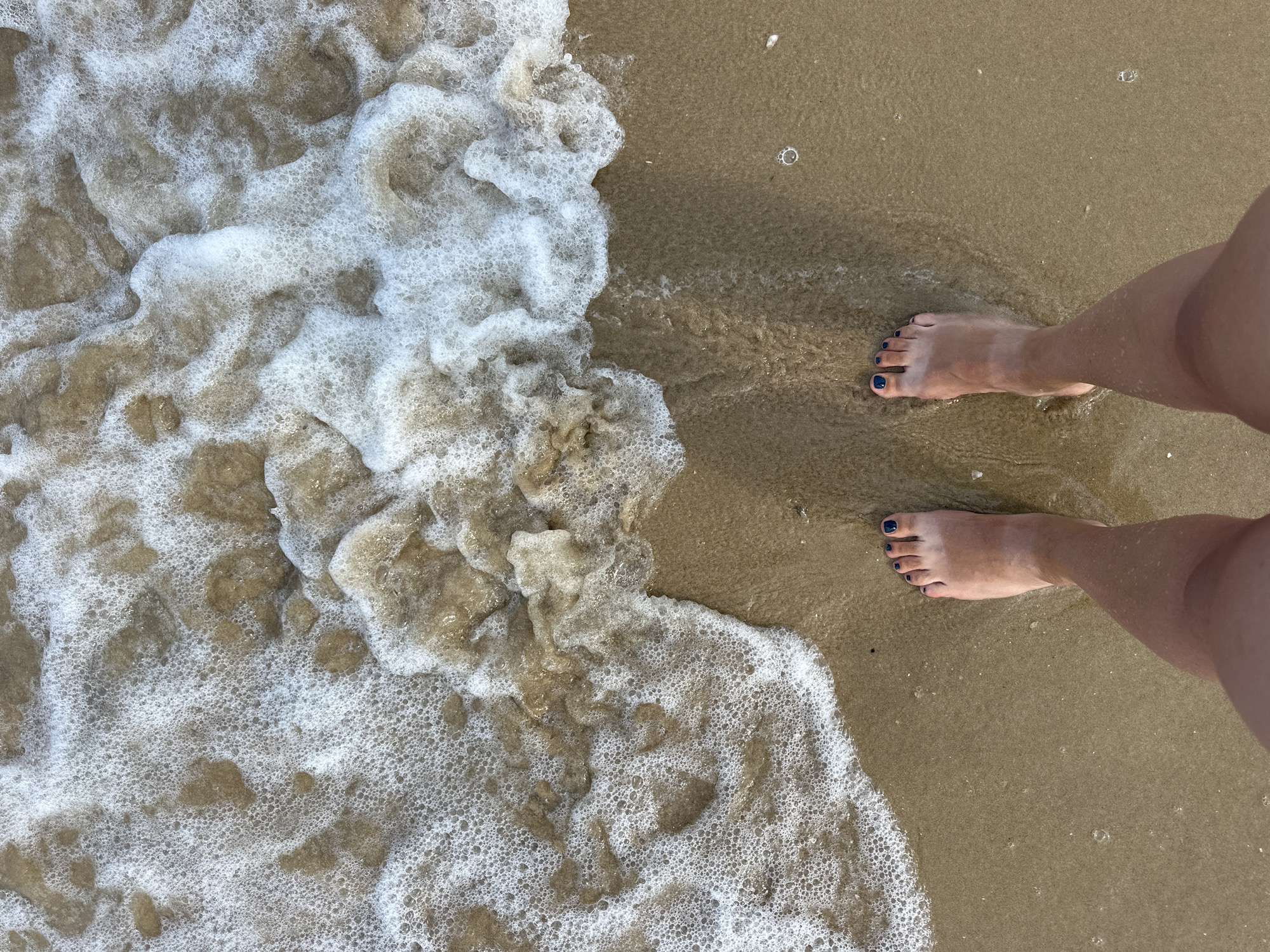 The image shows a pair of bare feet standing on a sandy beach. The feet are positioned in the lower right quadrant of the frame. The left foot is closer to the edge of the frame and is slightly submerged in foamy water. The right foot is further up the beach, in drier sand. Both feet have dark blue nail polish. The background is a sandy beach with the ocean waves and foam encroaching on the sand. The lighting suggests it is daytime. The overall scene depicts a person enjoying a moment at the beach.
