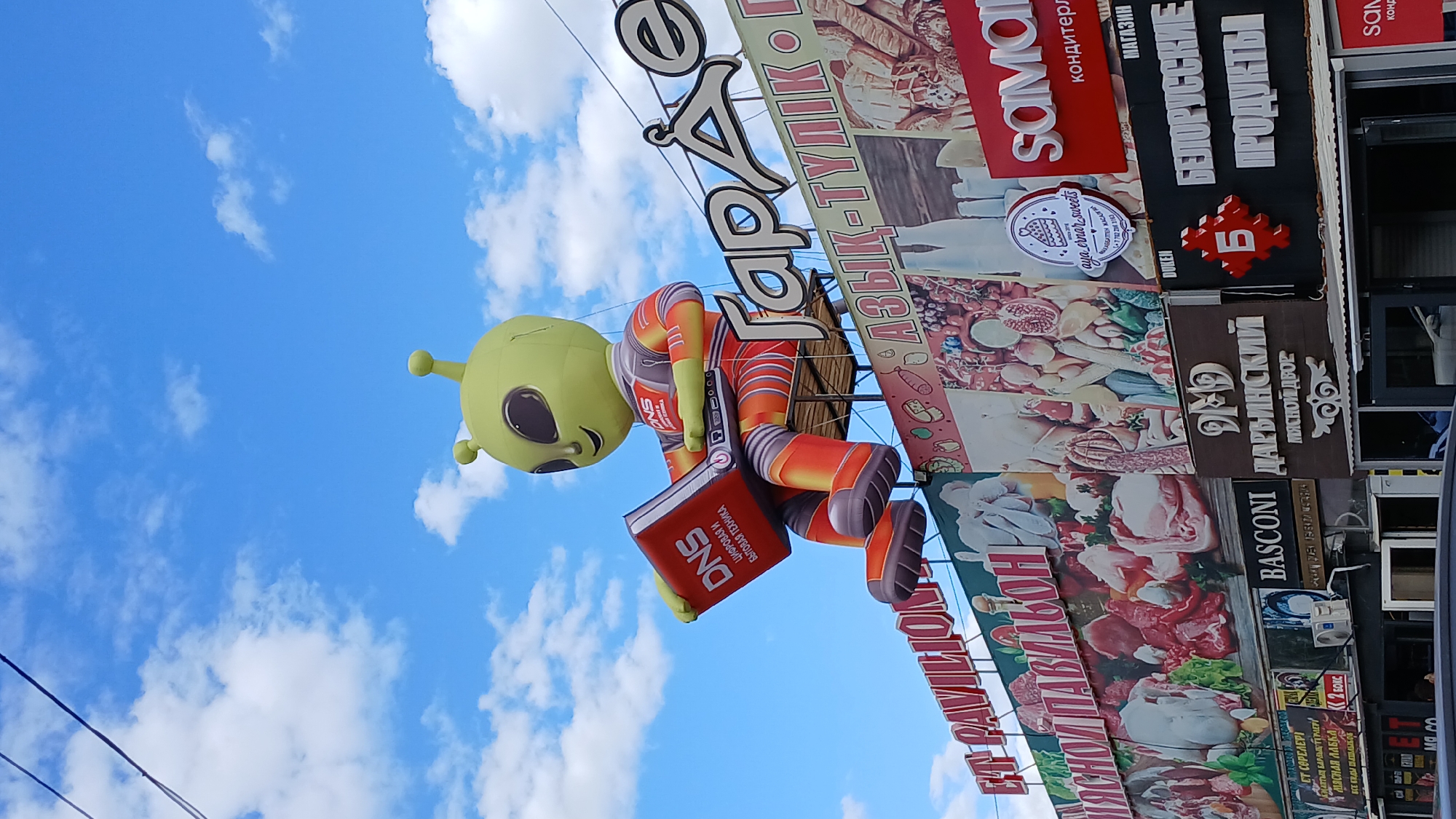 This is a low-angle, medium shot of an inflatable alien character holding an orange laptop, positioned in front of a large billboard featuring images of food products. The alien is bright green with large black eyes and antennae, wearing a futuristic orange and red suit. The laptop has the "DNS" logo and Russian text, likely indicating a technology store.
Behind the alien, a large sign in white, stylized Cyrillic letters reads "Гараж" (Garage). Below this, a billboard displays various food items, including meat, fruits, vegetables, and baked goods. Smaller signs are visible on the buildings below, with text such as "МЯСНОЙ ПАВИЛЬОН" (Meat Pavilion) and "ДАРВИНСКИЙ МЯСНОЙ ДВОР" (Darwin Meat Yard). Another sign reads "БЕЛОРУССКИЕ ПРОДУКТЫ" (Belarusian Products).
The scene appears to be a commercial area, possibly a market or shopping center, with multiple businesses advertised. The sky above is blue with scattered white clouds. The presence of a "DNS" logo and the style of advertising suggest this location is in a city where Russian is commonly used. Based on the Cyrillic text and the style of the businesses, it is highly probable that this image was taken in the city of Oral, Kazakhstan, which has a significant Russian-speaking population and a strong cultural connection to Russia. There are no people clearly visible in the image.