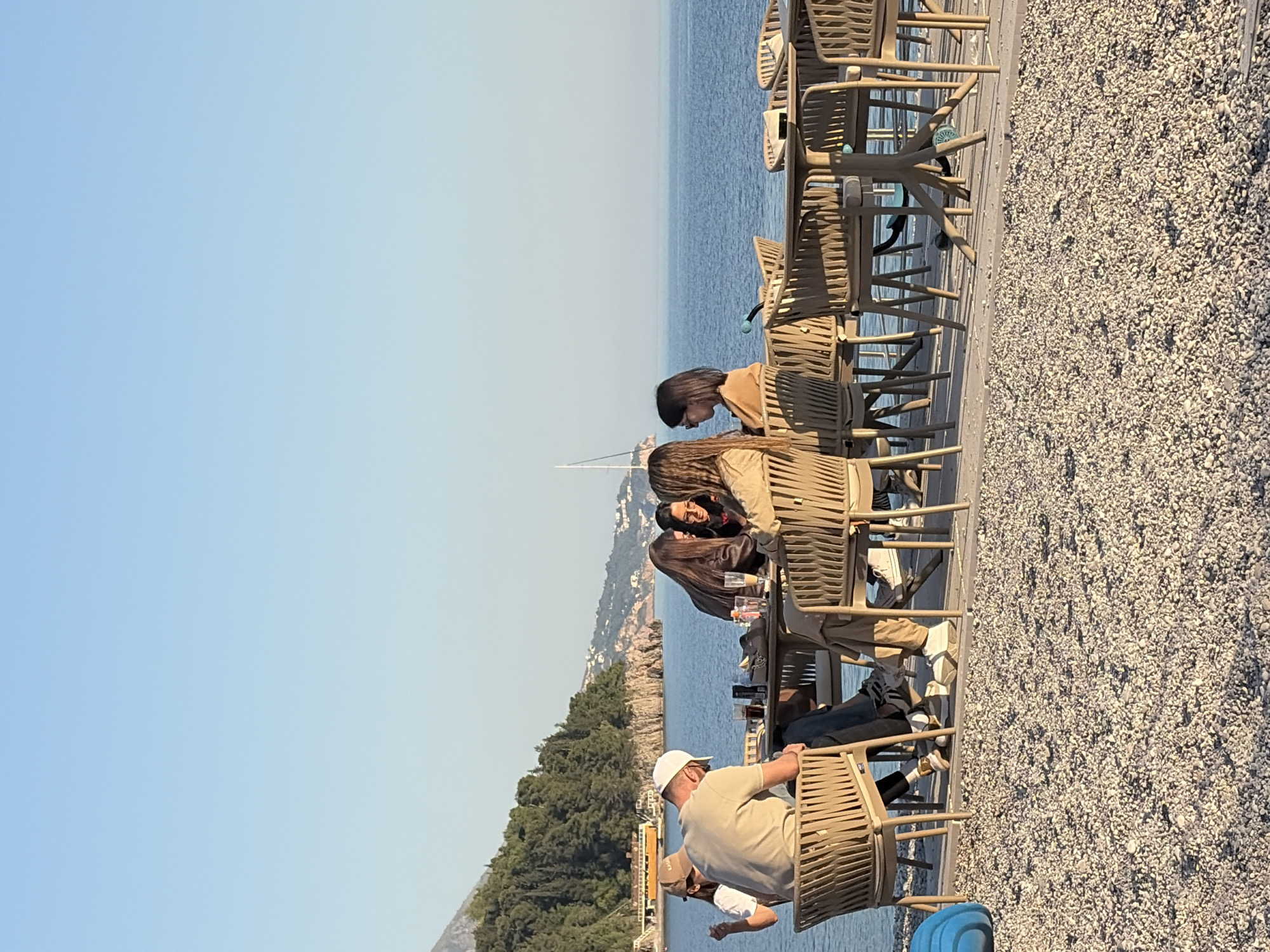 The image depicts a group of people seated at tables with woven chairs outdoors, possibly at a cafe or restaurant by the sea. The location is near a coastline with vegetation-covered cliffs and distant buildings. The sky is clear and blue. A pebbled surface covers the area to the right of the tables.