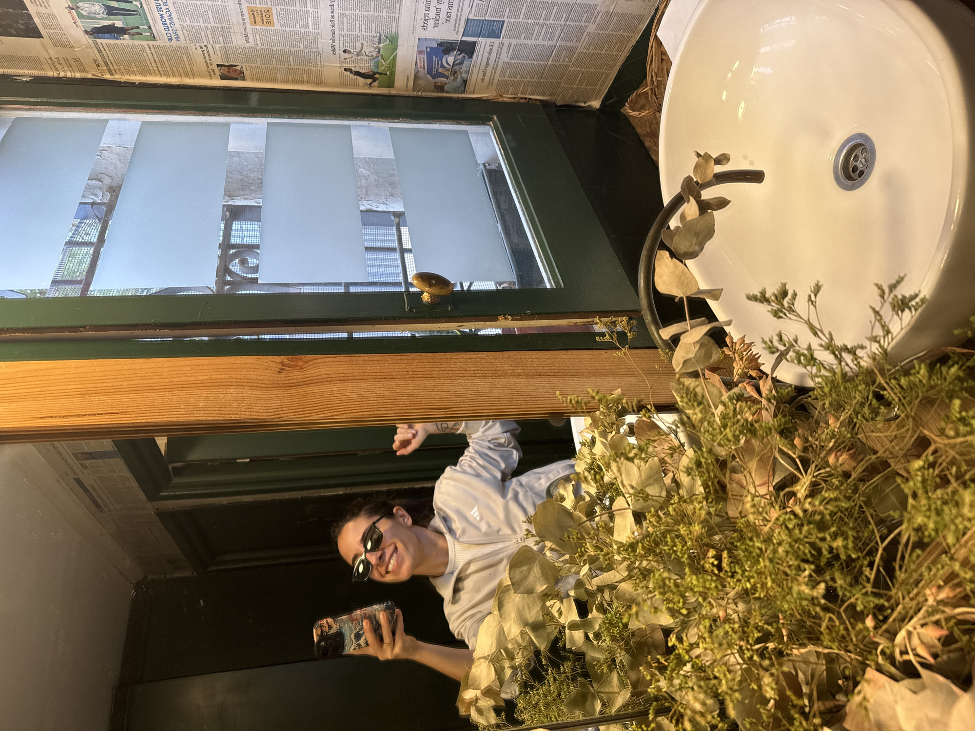 The image displays a woman taking a mirror selfie inside a distinct bathroom in Barcelona, Spain. The woman, appearing to be in her 20s or 30s, is reflected smiling broadly, wearing dark sunglasses and a light-colored Adidas zip-up jacket over a white top. She holds a smartphone with a decorative case in her left hand, while her right arm is raised in a fist-pump gesture, revealing a tattoo on her forearm.
The immediate foreground is filled with dried botanical arrangements, including fine green sprigs and larger, lighter-toned leaves, partially obscuring the lower section of the woman's reflection. Below the dried plants, a white, oval-shaped basin sink is visible, featuring a dark, curved minimalist faucet.
To the right of the mirror, a dark green-framed window is fitted with three horizontal strips of frosted film for privacy. Through the clear sections of the window, a view of exterior stone or stucco buildings and metal railings is discernible. The wall to the right of the window is covered with newspaper pages, prominently displaying text and images. One visible headline distinctly reads "LA SELECCIÓ CATALANA," indicating the location is within Catalonia. Dark green walls frame the mirror and window, and a vertical wooden beam separates the two. A woven basket is partially visible on the floor or counter beneath the newspaper-covered wall.