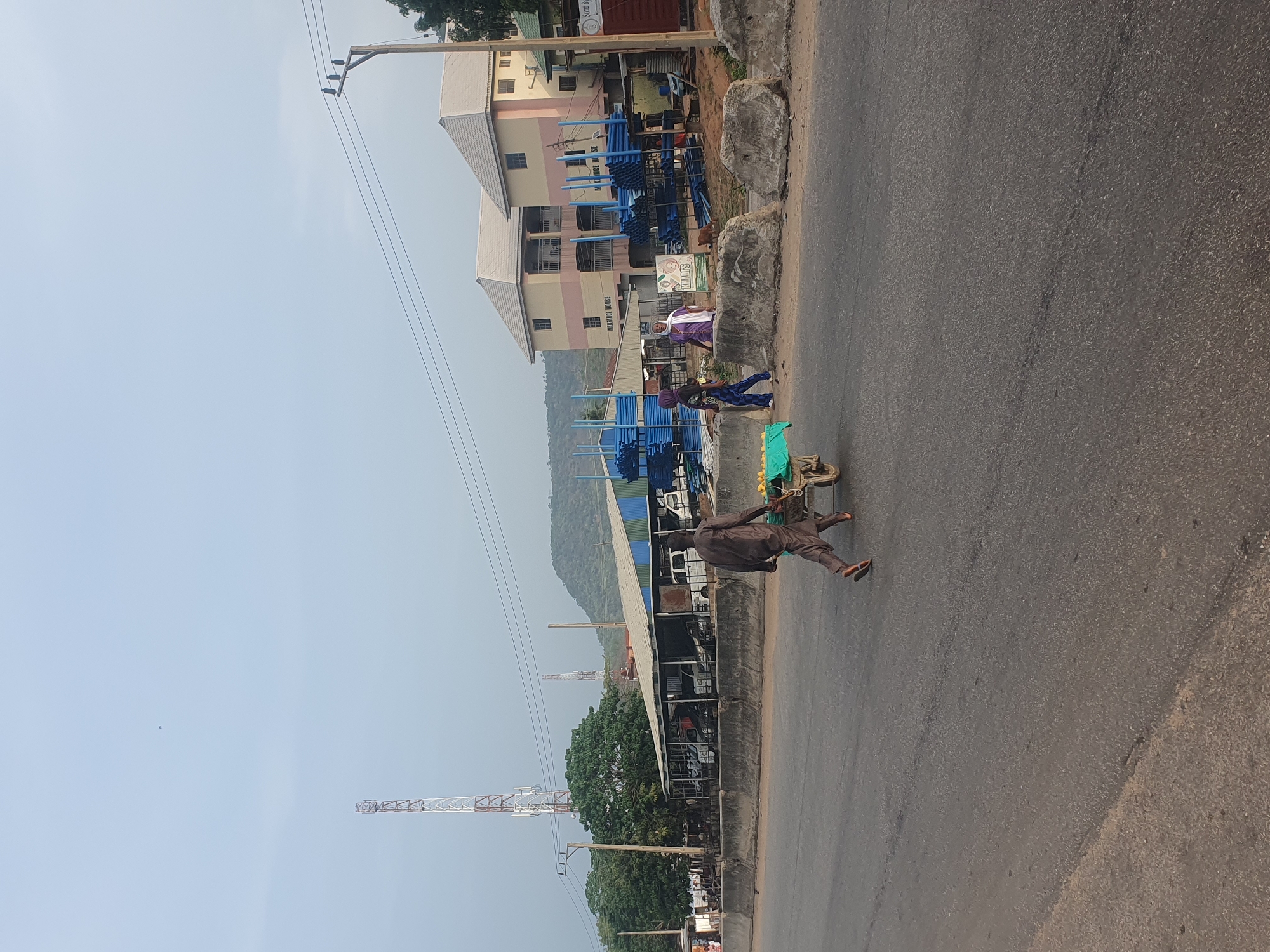 A medium, slightly high-angle shot, taken in daylight, reveals a man pushing a wheelbarrow laden with green objects along the side of a street in Lokoja, Nigeria. The man is wearing brown clothing and sandals. A building of two-stories is present, with stacked blue pipes outside of it. Adjacent to the building is a corrugated metal structure, and the area is near the edge of town. A tall radio tower rises in the background. A woman in a purple and white outfit stands by the blue pipes. Other people can be seen in the area.