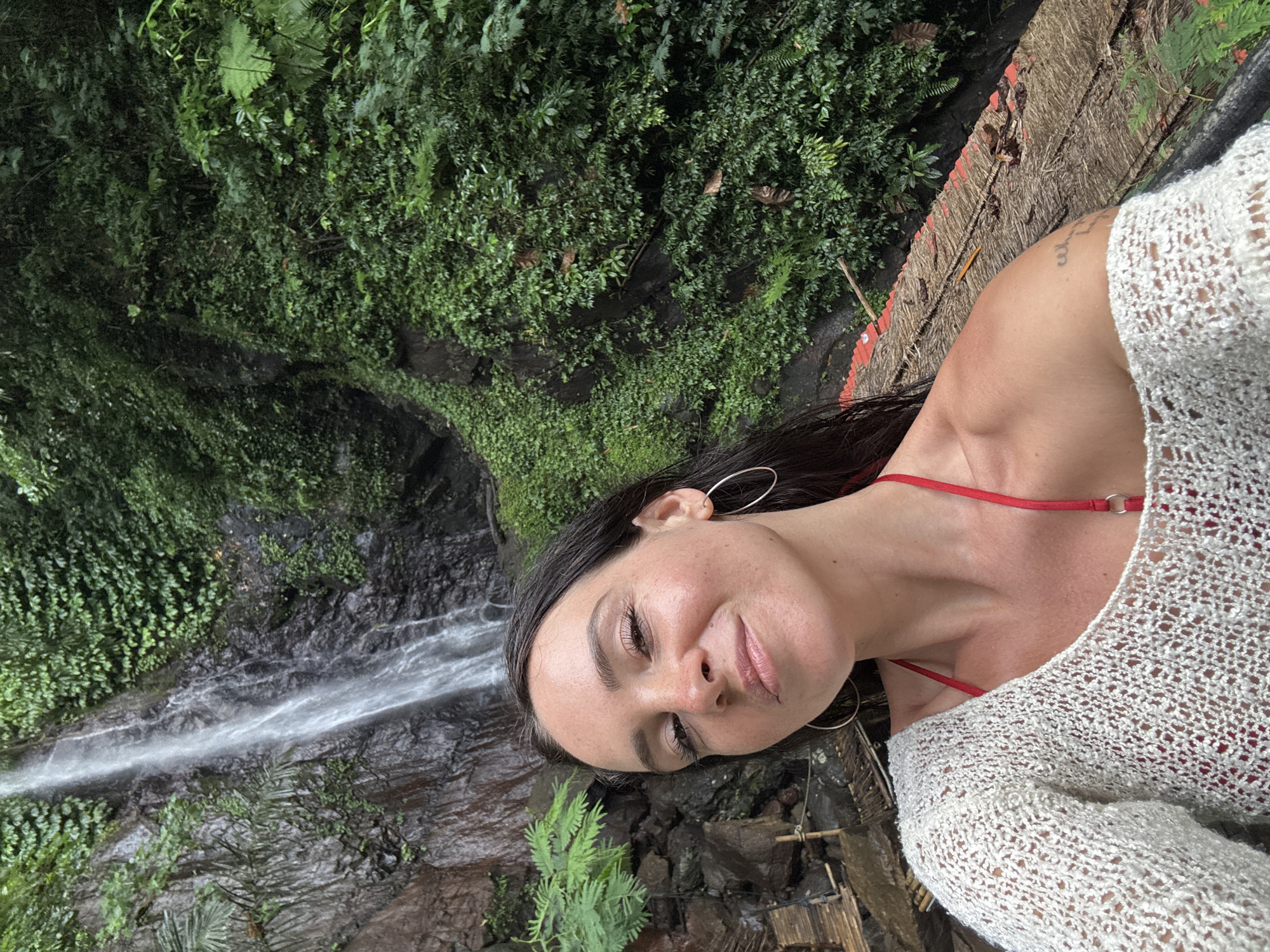 A woman with dark hair, wearing a white crocheted top over a red bikini and large hoop earrings, takes a selfie in front of a waterfall. The waterfall cascades down a dark, rocky cliff face covered in lush green foliage. The woman is positioned in the foreground, smiling slightly. To the left of the waterfall, a wooden structure with bamboo railings is visible, partially obscured by greenery. The overall environment is a tropical jungle. The image appears to be taken in or near Selat, Indonesia, given the natural landscape and vegetation commonly found in the region.