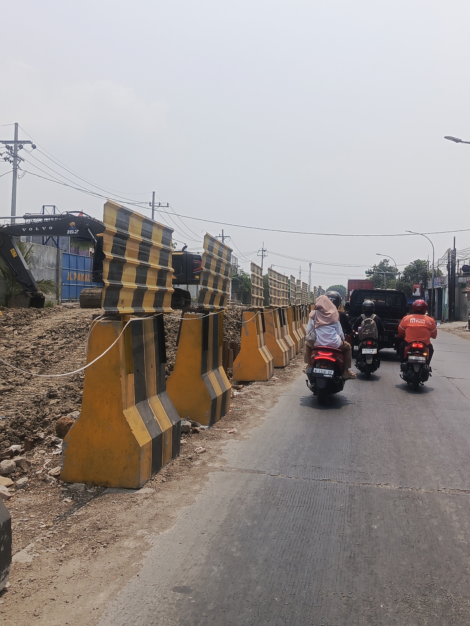 The image depicts a street scene in Surabaya, Indonesia, characterized by ongoing road construction and traffic. In the foreground, concrete barriers with black and yellow stripes cordon off a construction zone on the left side of the road. An excavator, bearing the "VOLVO" logo and the number "162," is visible within the construction area. On the right side of the road, several motorcycles are in motion. One motorcycle carries two passengers, the rear passenger wearing a light brown hijab and white top, and the driver wearing a dark helmet. Another motorcycle, further ahead, has a driver with a backpack, and a third motorcycle is ridden by a person in an orange uniform with "Food" written on the back, suggesting a delivery service. A black truck is positioned behind the motorcycles. The environment is urban, with overhead power lines and streetlights present. The sky is overcast, indicating a possibly hazy or cloudy day. The road surface shows signs of wear and tear.