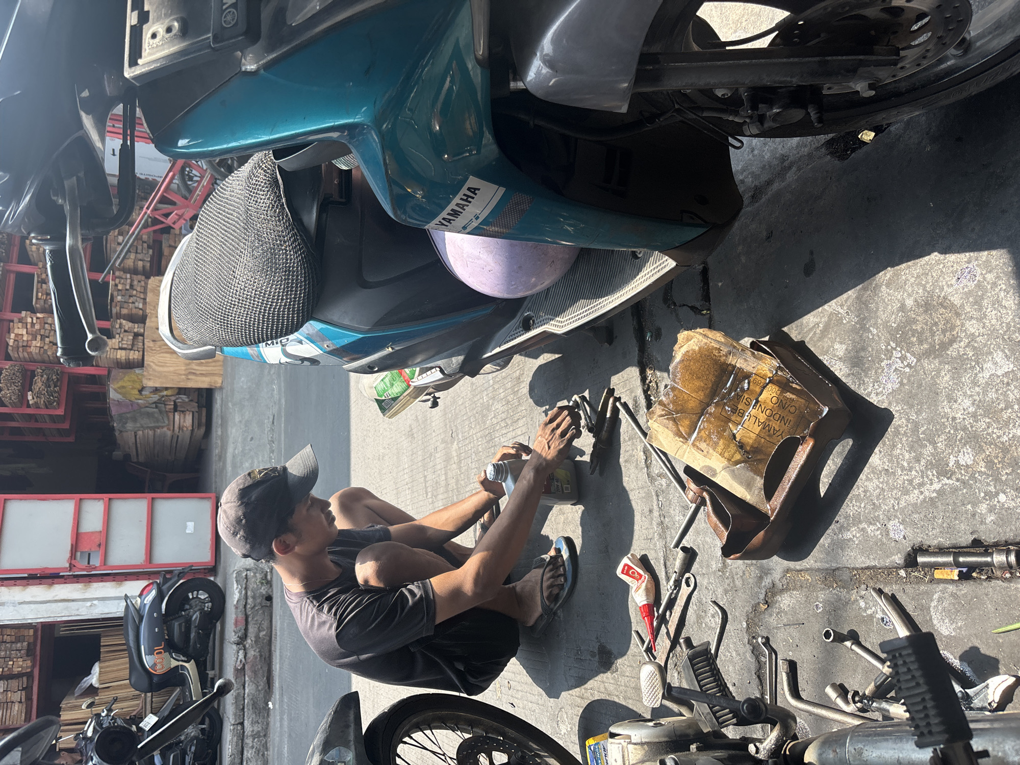 In North Jakarta, Indonesia, a young man wearing a dark t-shirt and a black baseball cap squats on the pavement, appearing to perform maintenance on a teal scooter. He is holding a bottle of what appears to be oil in his right hand and a tool in his left. Various tools and motorcycle parts are scattered on the ground around him, including wrenches, a brush, and a metal container filled with oily rags. In the background, a store with stacks of wood and a red-framed door is visible, suggesting an industrial or market area. Another parked scooter is also visible to the left of the frame. The scene is bathed in harsh sunlight, casting distinct shadows on the concrete ground.