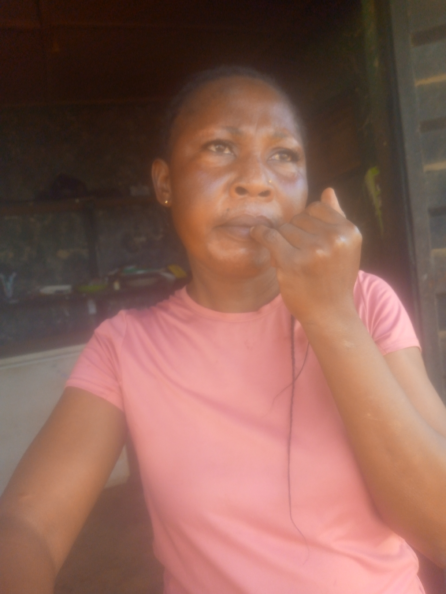 The image features a close-up portrait of a person, likely a woman, with dark skin and a thoughtful or pensive expression. She is captured from the chest up, looking upwards and slightly to her right. Her right hand is raised, with her index and middle fingers gently touching her lower lip.
She is wearing a plain, short-sleeved pink t-shirt. Her hair appears to be pulled back, and a single strand of dark hair hangs down in front of her right shoulder. A small, gold-colored stud earring is visible in her right ear. Notable skin characteristics include several lighter patches on her forehead, chin, and the back of her right hand, suggesting variations in skin pigmentation.
The background is dark and somewhat blurry, suggesting an indoor or heavily shaded outdoor setting. On the left side of the frame, indistinct shapes can be seen, possibly shelves or furniture against a dark wall. A vertical dark line on the right side might indicate a doorframe or window frame. The lighting appears natural, illuminating her face from the front and slightly to the left. The overall mood conveyed is one of contemplation or deep thought. There is no visible text in the image.