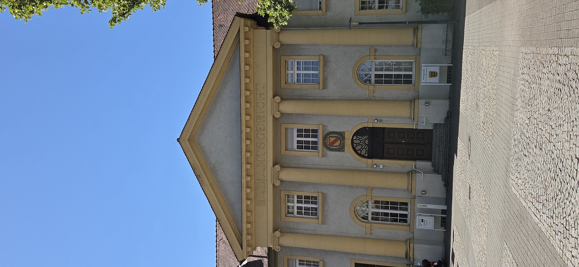 The image shows a frontal view of the Amtsgericht (District Court) in Kehl, Germany. The building has a classical facade with large yellow columns, a triangular pediment, and the inscription "BADAMTSGERICHT" above the entrance. The entrance itself features a large, dark wooden double door topped by a decorative arched window. Above the door is a coat of arms. The building is flanked by windows with white frames. The ground in front of the court is paved with cobblestones and some rectangular pavers. A sign in front of the building reads "Amtsgericht" with the coat of arms of Baden-Württemberg. To the left of the entrance, a motorcycle is parked. A tree with green foliage is visible on the right side of the image, and the sky is clear and blue.