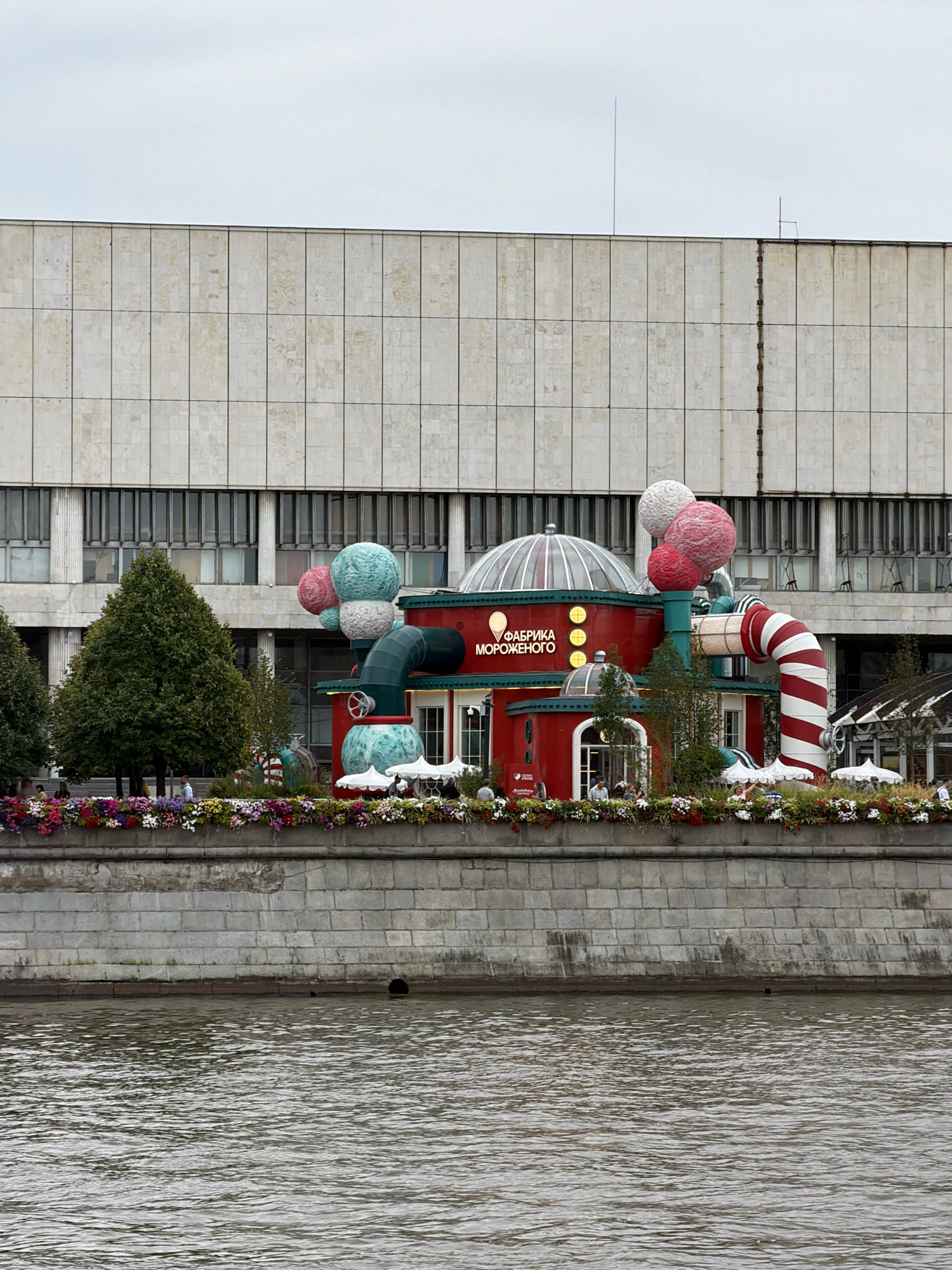 The image shows the "Ice Cream Factory" pavilion located on Muzeon Park of Arts in Moscow, Russia, situated along the Moskva River. The pavilion is a brightly colored, whimsical structure with a red main body, a glass dome, and large, spherical decorations resembling ice cream scoops. A large, red and white striped pipe extends from the structure. The pavilion is adorned with an abundance of colorful flowers along its waterfront edge. Behind the pavilion, a large, imposing building with a concrete facade and numerous windows can be seen. The Moskva River occupies the foreground, with ripples indicating a slight current. Several people are visible, some near the pavilion and others further back in the park, suggesting a public space. The sky is overcast and gray.