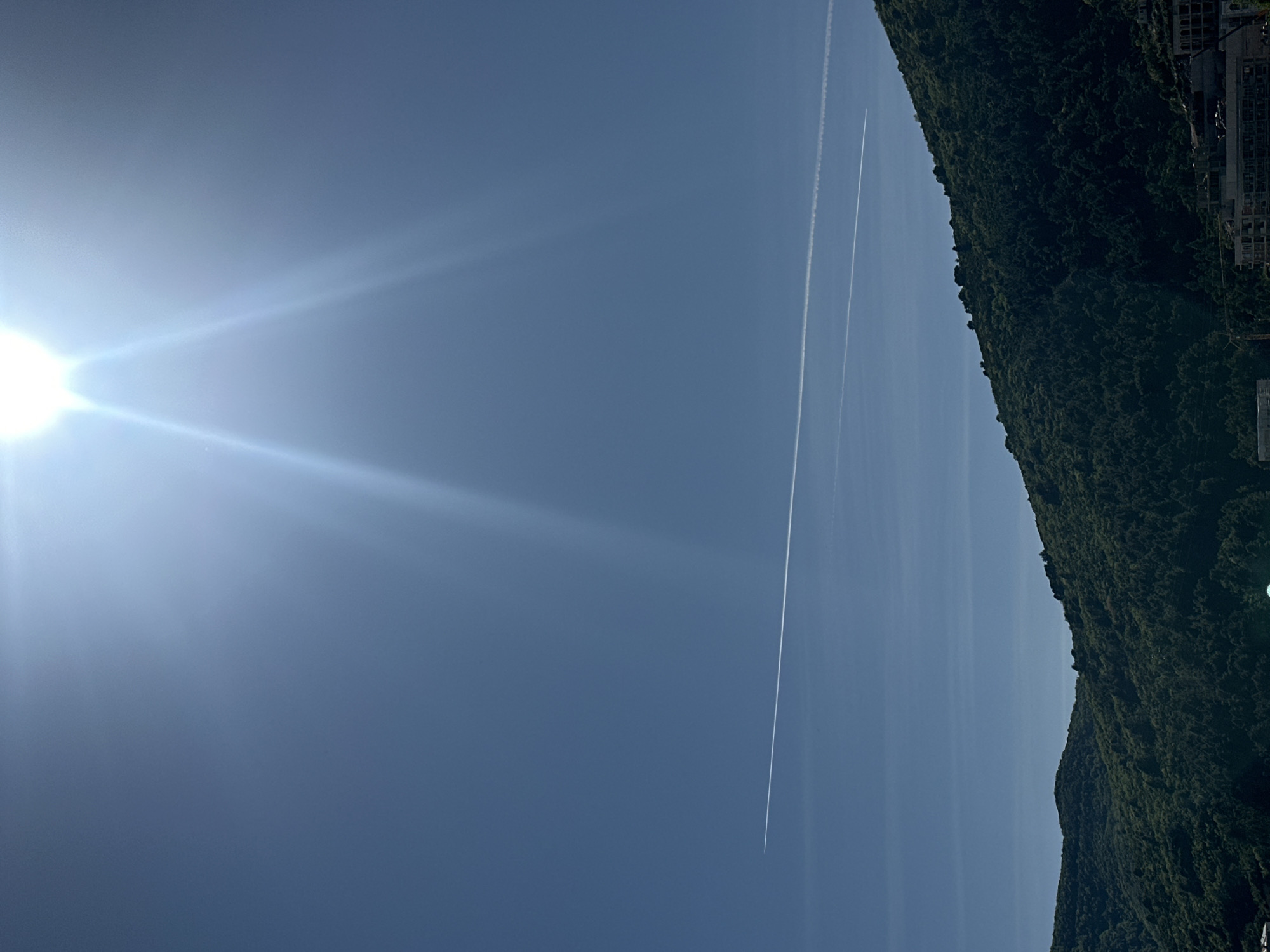 The image captures a bright, sunny daytime scene dominated by a clear blue sky and a verdant mountainous landscape. The sun, a brilliant white light source, is prominently featured in the upper left quadrant, casting strong lens flares and bright rays across the sky.
Two distinct, thin white contrails (vapor trails from aircraft) streak across the sky, running roughly parallel to each other from the upper right toward the lower left of the frame. These trails are the only visible elements in the otherwise clear expanse of the sky.
Along the right side of the image, a lush, dark green, densely forested mountain or hillside rises, filling approximately one-third of the frame. The varied textures of the tree canopy are visible. Towards the top right edge of the mountain, some light-colored man-made structures, possibly buildings or a development, can be seen nestled among the trees.
The overall impression is one of a clear, warm day. Given the context, this could be a view overlooking the scenic coastline and mountains of Budva, Montenegro. There is no visible text in the image.
