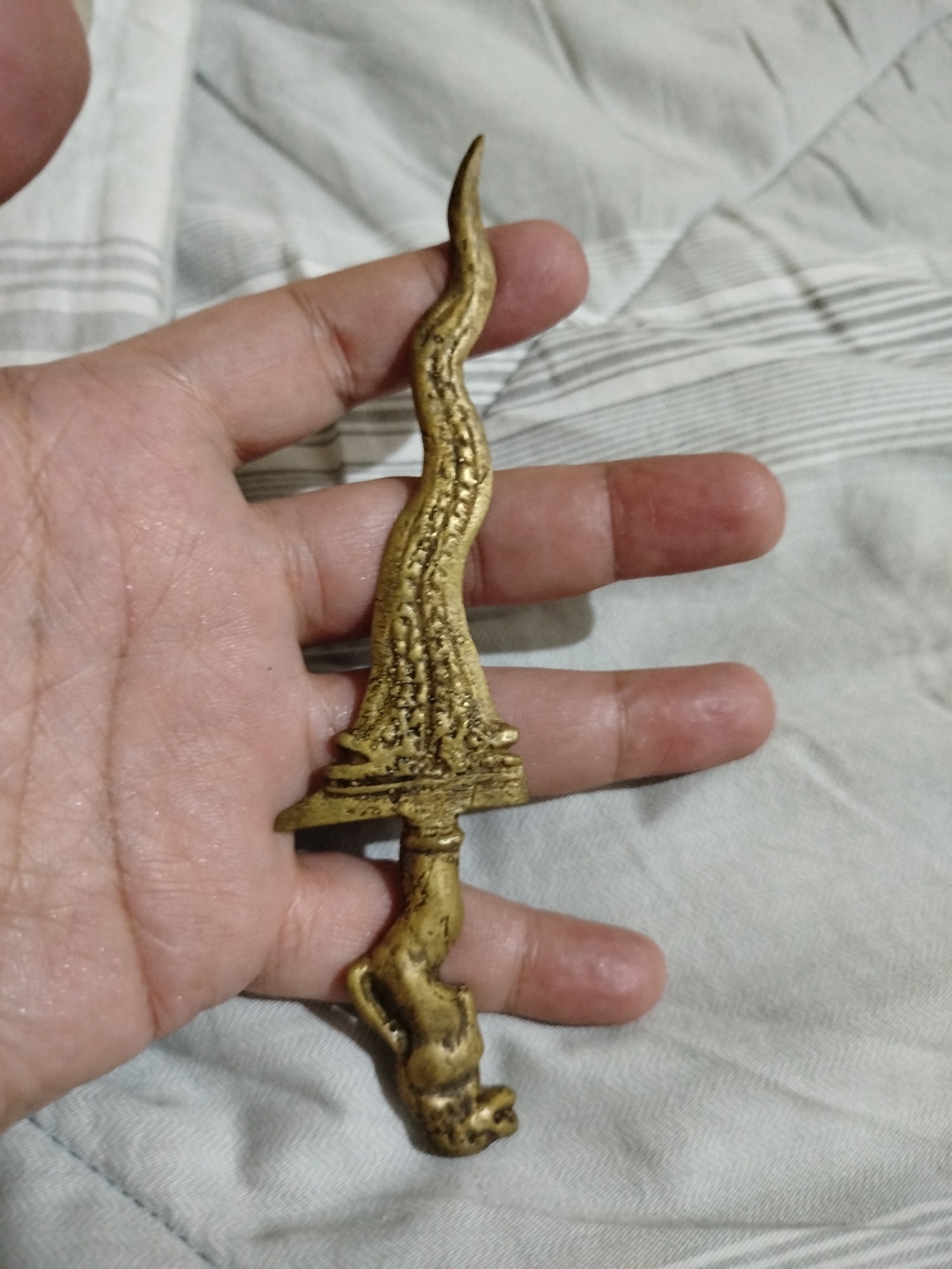 A hand is holding a small, ornate golden dagger. The dagger has a wavy blade and a hilt that resembles a seated animal figure. The background is a light-colored, textured fabric. The location is not specified within the image.