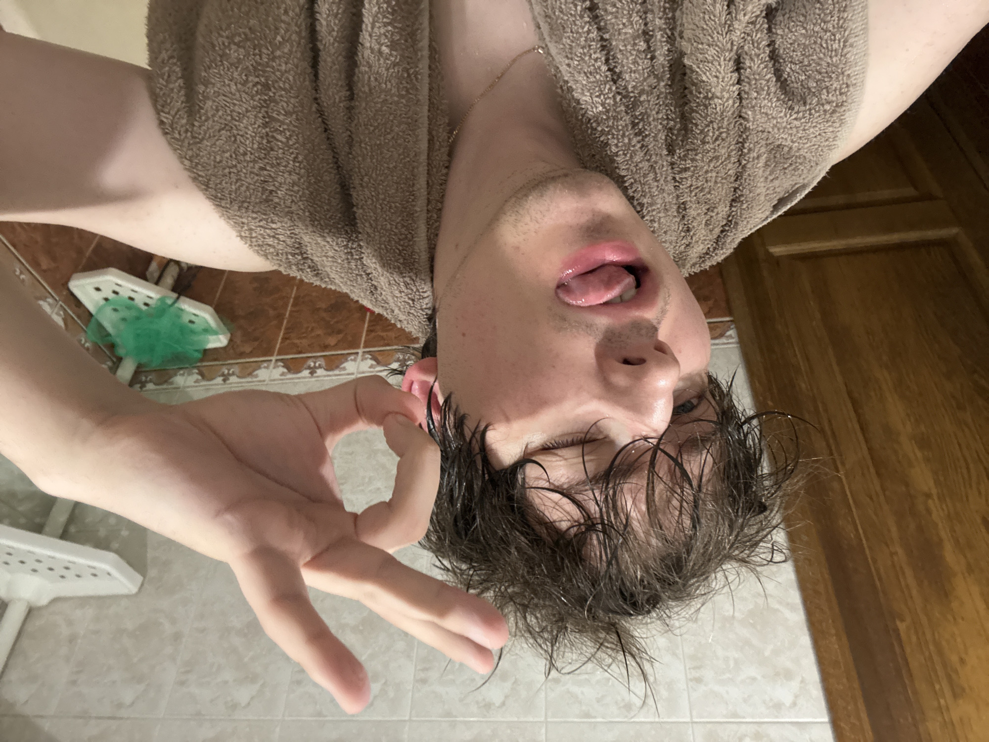 In a bathroom in Dnipro, Ukraine, a young man with wet, messy hair is taking a selfie. He is wearing a brown towel around his shoulders and a gold chain necklace. He is sticking out his tongue and winking at the camera, with his right hand making an "ok" sign near his face. The bathroom has tiled walls and a wooden door is visible on the left. A white shower caddy with a green sponge is mounted on the wall in the background.