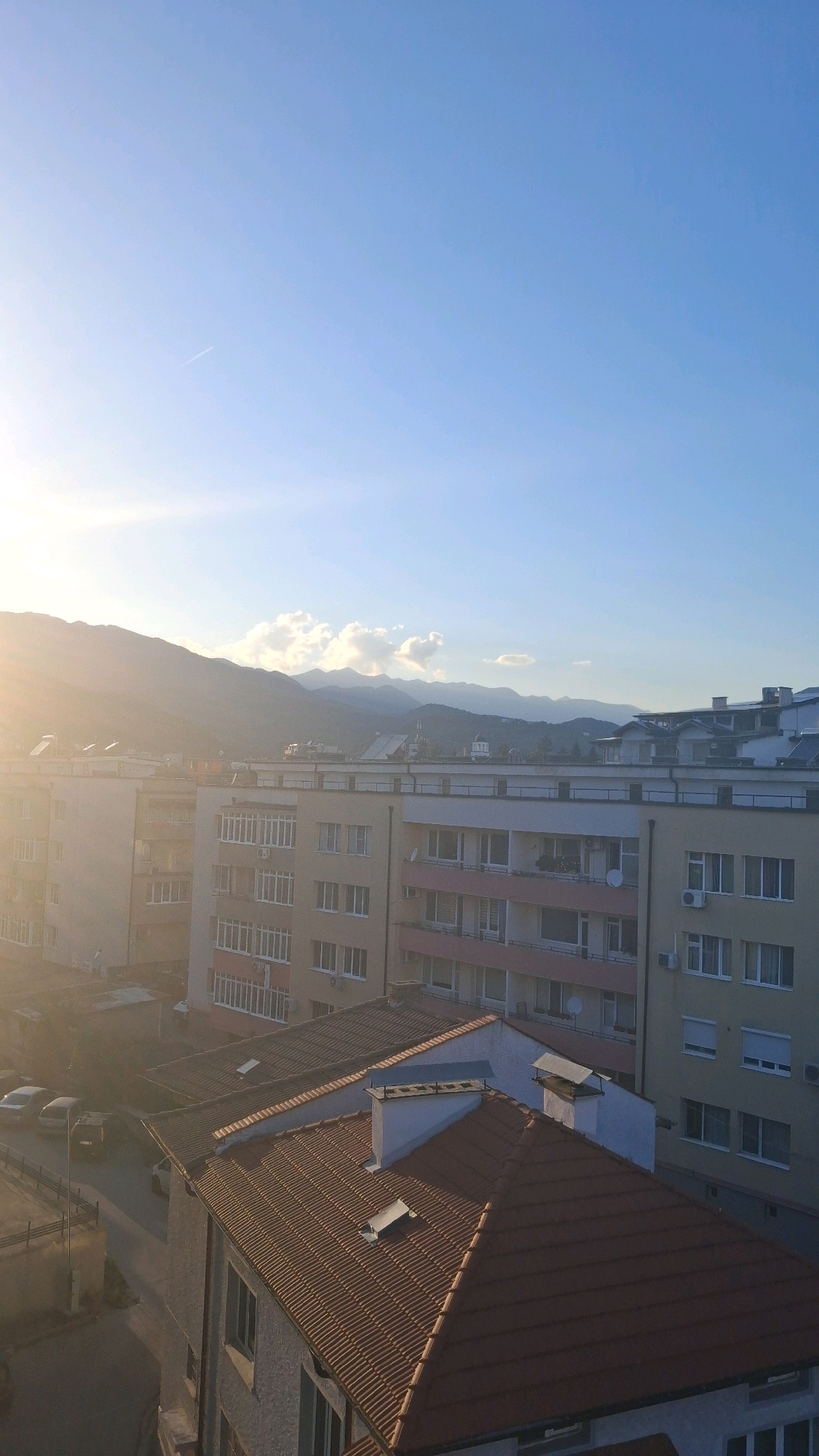 The image shows a view of the city of Gotse Delchev, Bulgaria, from a high vantage point. The foreground is dominated by the tiled roofs of residential buildings, with chimneys and ventilation systems visible. In the middle ground, several apartment buildings with multiple stories and balconies are present. The buildings are of various colors, including beige, yellow, and pink accents. Some windows are open, and air conditioning units are mounted on some facades. The background reveals a range of mountains under a clear blue sky. The sun is setting or rising, casting a warm, hazy glow on the scene and creating lens flare in the upper left portion of the image. A faint contrail from an airplane is visible in the sky. Several parked cars can be seen on a street in the lower left of the frame. The overall atmosphere suggests a quiet urban residential area during either dawn or dusk.