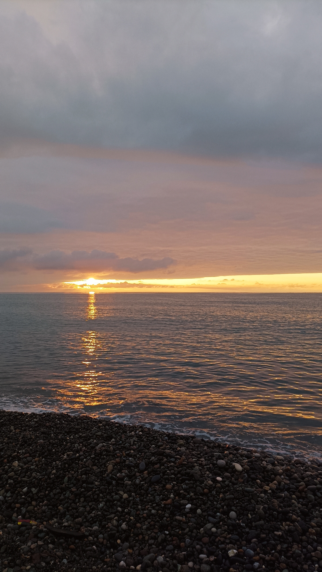 The image depicts a coastal scene at twilight, likely sunset, on the Black Sea coast in Batumi, Georgia. The upper portion of the frame is occupied by an overcast sky, displaying gradients of dull grey and light blue-grey clouds. Towards the horizon, a horizontal band of clear, warm light transitions from soft pink and orange to bright golden yellow, indicating the position of the sun. The sun itself is partially obscured by low-lying clouds, appearing as a brilliant golden point source.
The middle ground consists of the Black Sea, which has a dark, deep blue-grey surface marked by ripples and gentle waves. A prominent, shimmering vertical column of intense golden-orange light reflects off the water, extending from the sun towards the foreground.
The foreground is a pebble beach composed of numerous dark, smooth, rounded stones of varying sizes. The water gently laps at the shore, creating a wet line on the pebbles with minimal white foam from small waves. No individuals, animals, structures, or man-made objects are visible within the frame. The scene is unpopulated, depicting a natural coastal environment.