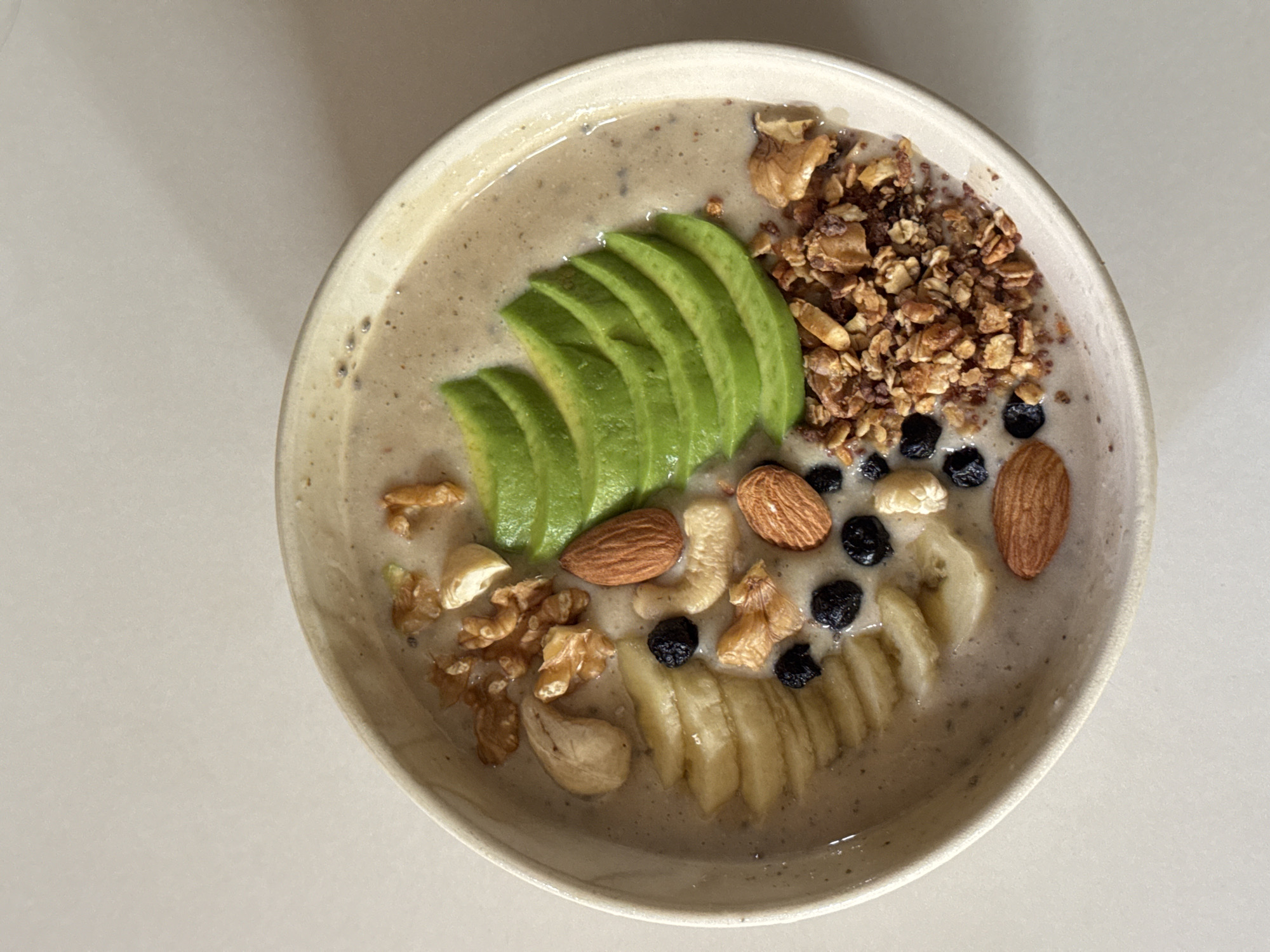 A high-angle, medium shot presents a bowl of smoothie in a ceramic bowl. The smoothie is decorated with sliced avocado, banana, granola, blueberries, walnuts, cashews, and almonds. The background is a plain, light-colored surface.
