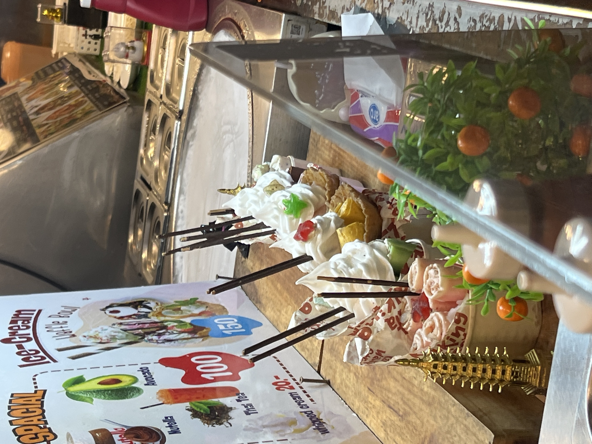 This media file shows a close-up of an ice cream display at a food stall, likely in Karon, Thailand. The focus is on various ice cream desserts presented in waffle bowls and paper cups, adorned with whipped cream, fruit toppings like cherries and what appear to be small tangerines on a decorative plant, and chocolate sticks. Some of the desserts are being served with individual brown paper wrappers that have red and white text.
Prominently displayed in the foreground is a menu board with images of food items and text. Visible text on the menu includes "Ice-cream Waffle Bowl", "Special", "Avocado", "Nutella", "Thai Tea", "Whipped Cream 20-", and prices "150" and "100". The overall scene is vibrant and suggests a busy, open-air food environment, typical of a tourist area. The lighting indicates it is daytime. The display is set up behind a glass barrier, implying it's a service counter. The decorative elements, like the tiny golden pagoda-like ornament, add to the local flavor of the setting.