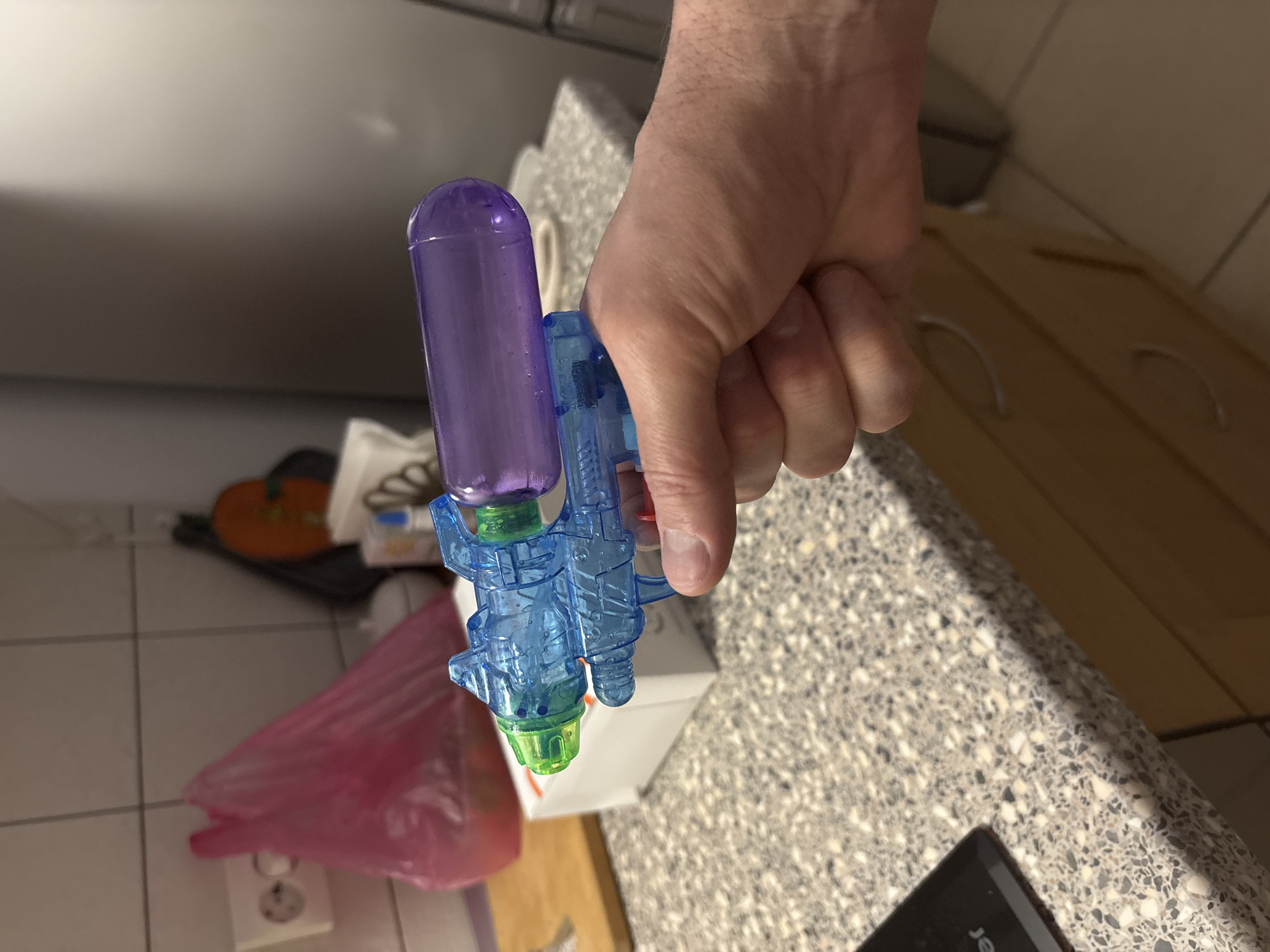 A person holds a small, colorful plastic water gun. The gun is primarily blue, with a purple reservoir and green accents. The scene appears to be indoors, possibly in a kitchen, with a countertop and tiled walls visible. A pink plastic bag and other household items are scattered in the background. The location is not identifiable as Saint Petersburg, Russia, based solely on the image.