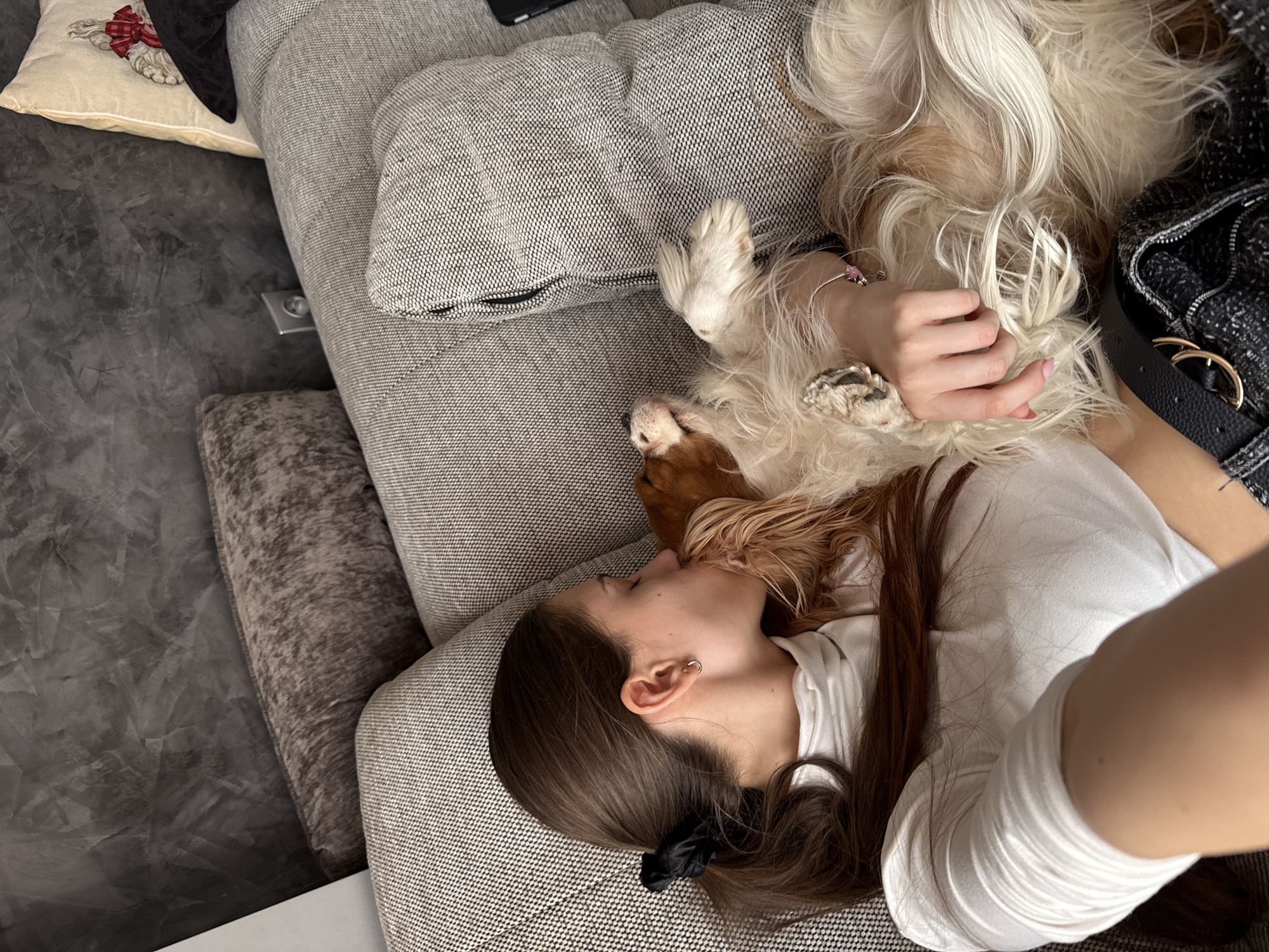 A high-angle shot depicts a woman reclining on a light gray couch indoors. She is wearing a white t-shirt, and her long brown hair is partly tied back with a black scrunchie. She's lying on her side, her face close to a Cavalier King Charles Spaniel dog. The dog is lying upside down, with its paws near the woman's face. The dog is a mix of white and brown fur. The woman is holding the dog. A black and white patterned handbag with a black leather belt sits nearby on the couch. A throw pillow with an image of a bow and two other cushions are on the couch. The floor is a dark gray polished concrete. There is no evidence to suggest that the location is in Sofia, Bulgaria.