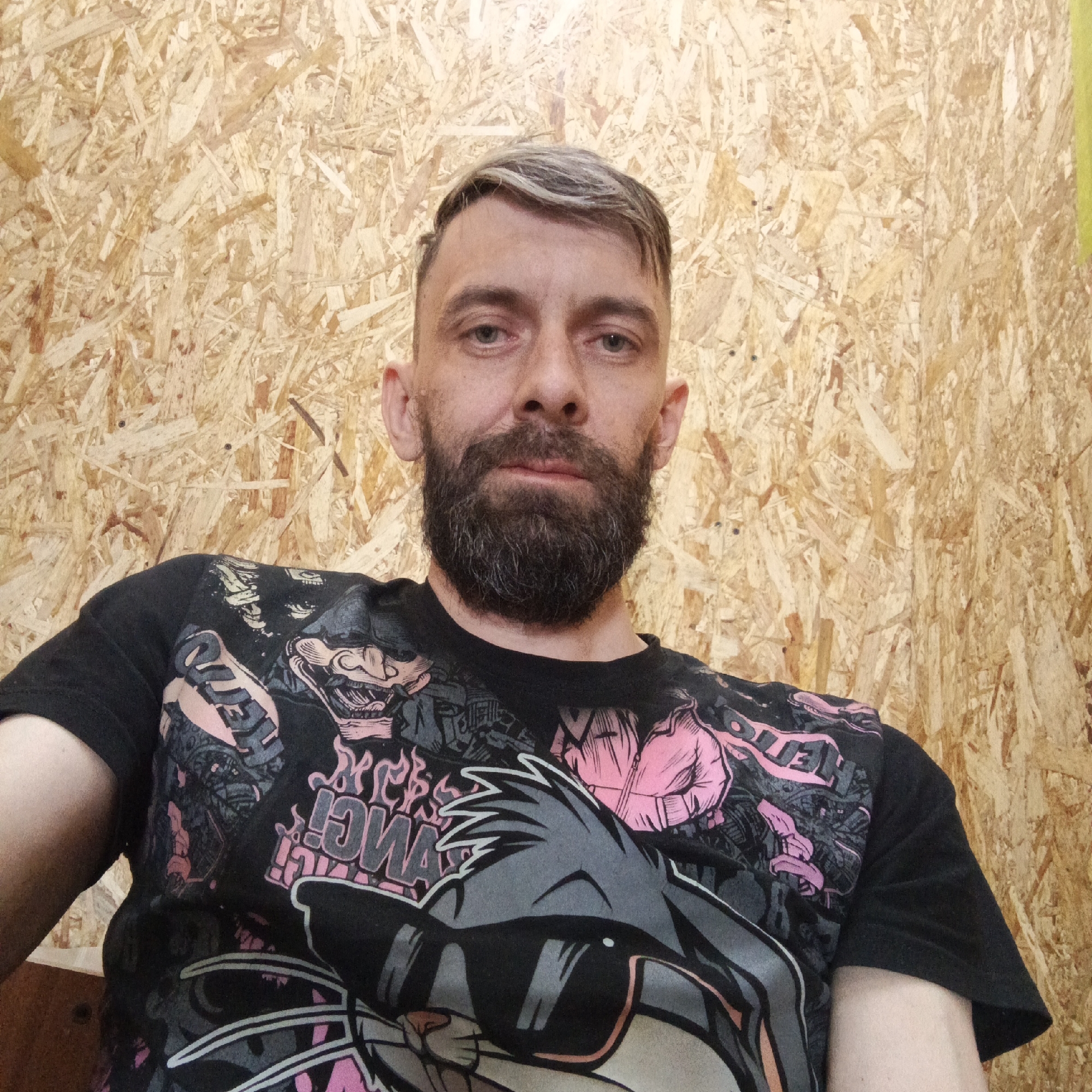 A medium-shot self-portrait captures a Caucasian male with a fair complexion, light gray-streaked hair, and a full, dark beard. He is situated indoors, against a rough, composite wood wall. The subject is wearing a black t-shirt printed with cartoon figures in black, gray, and pink hues.
The location is identified as Chaikovsky, Russia.