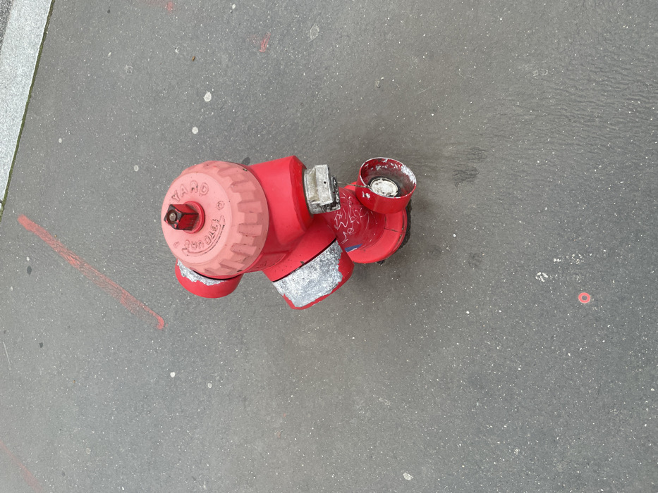 The image displays an overhead view of a red fire hydrant positioned on a dark gray asphalt surface. The hydrant features a ridged, pinkish-red top cap inscribed with the text "YACO," "100," and "5 3/4 TG 1978," and is equipped with a square operating nut. The main body of the hydrant is red, exhibiting areas of worn silver-colored paint or material around its upper and mid-sections. White graffiti is visible on the lower red portion of the hydrant. Two outlets are present; one is a larger, capped port, while the other is a smaller, uncapped port with a white interior, showing wear and exposure. The surrounding asphalt pavement contains a distinct diagonal red line marking in the upper left quadrant and a faint small red circular marking near the lower left. White specks are distributed across the asphalt, which also shows dark, irregular moisture or stain marks immediately around the base of the hydrant. A lighter gray concrete curb or sidewalk edge is visible along the upper right perimeter of the scene. This location is in Montigny-le-Bretonneux, France.