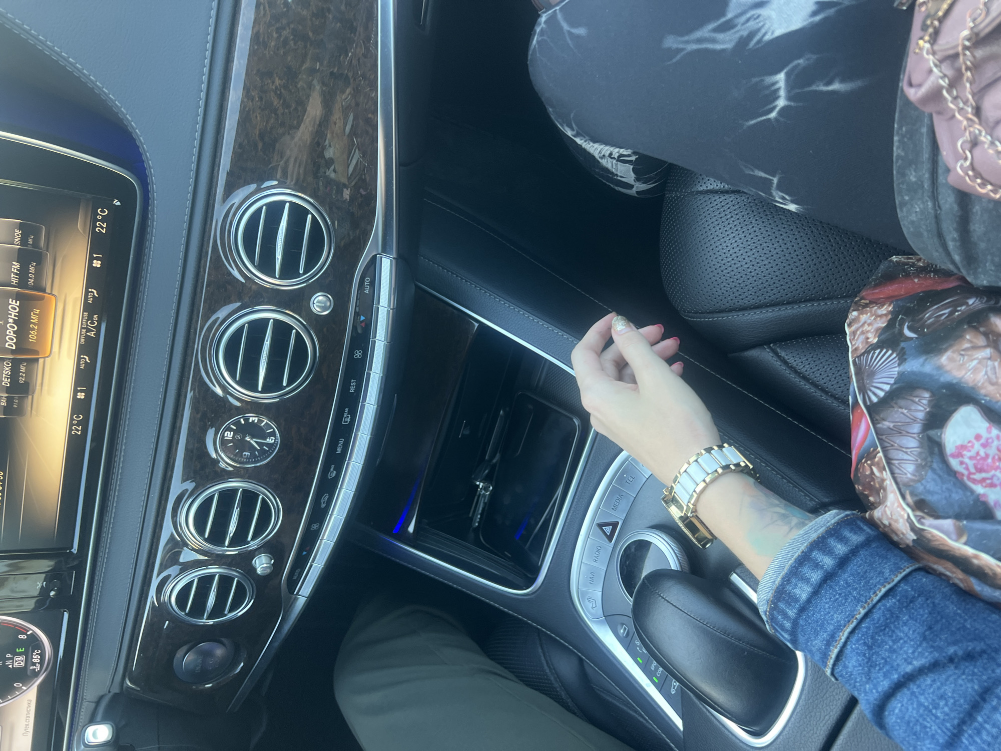 The image shows the interior of a car. In the foreground, a person's arm, wearing a denim jacket and a watch, is visible. The person's left hand is resting on the car's center console. In the background, the dashboard and car's entertainment system are visible. The car is likely parked in Krasnoe-na-Volge, Russia, given the radio station names visible on the display (e.g., "HIT FM," "SNOE," "DETS"KO"). The climate control system is set to 22°C.