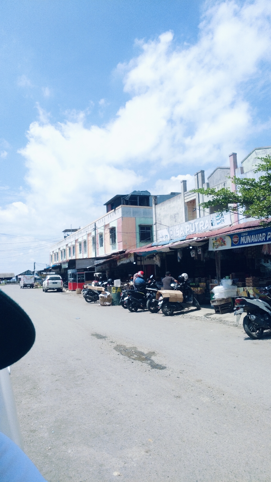 This is a daytime, eye-level, medium-wide shot of a street in Lhokseumawe, Indonesia, on a cloudy day. On the left side of the frame, a white car is parked. Further down the street, a white truck is visible. In the center of the frame, there are several motorcycles parked in front of shops. Some people are visible sitting on the motorcycles and in front of the shops. The shops appear to be selling various goods. The buildings are multi-story structures with shops on the ground floor. The sky is blue with white clouds. The road is paved with asphalt. There are also some trees visible on the right side of the frame.