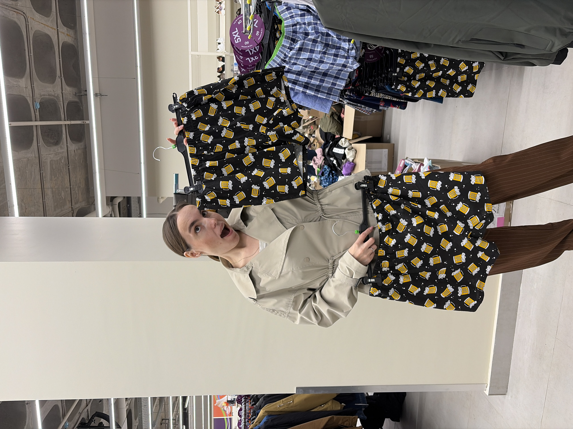 A young woman in a retail store in Izhevsk, Russia, holds up a pair of black boxer shorts decorated with a repeating pattern of yellow beer mugs, displaying an expression of surprise or excitement with her mouth open and eyes wide. She wears a light beige trench coat or jacket over a white top and brown pinstriped trousers.
The indoor setting appears to be a men's clothing section, possibly within a larger department or discount store. To her right, another identical pair of beer-themed boxer shorts hangs on a rack. The background is filled with various other clothing items, including blue plaid boxers, hanging on metal racks. Numerous cardboard boxes, some open and containing additional apparel, are visible on the floor, suggesting a stock or less organized area of the store. The ceiling is made of concrete with square recessed sections, illuminated by long fluorescent strip lights.
Visible text on round purple tags attached to clothing racks includes sizes "5XL" and "4XL," along with the Russian word "МУЖЧИНЫ" (MUZHCHINY), meaning "MEN," and the number "60." A white tag with the number "58" is also discernible.