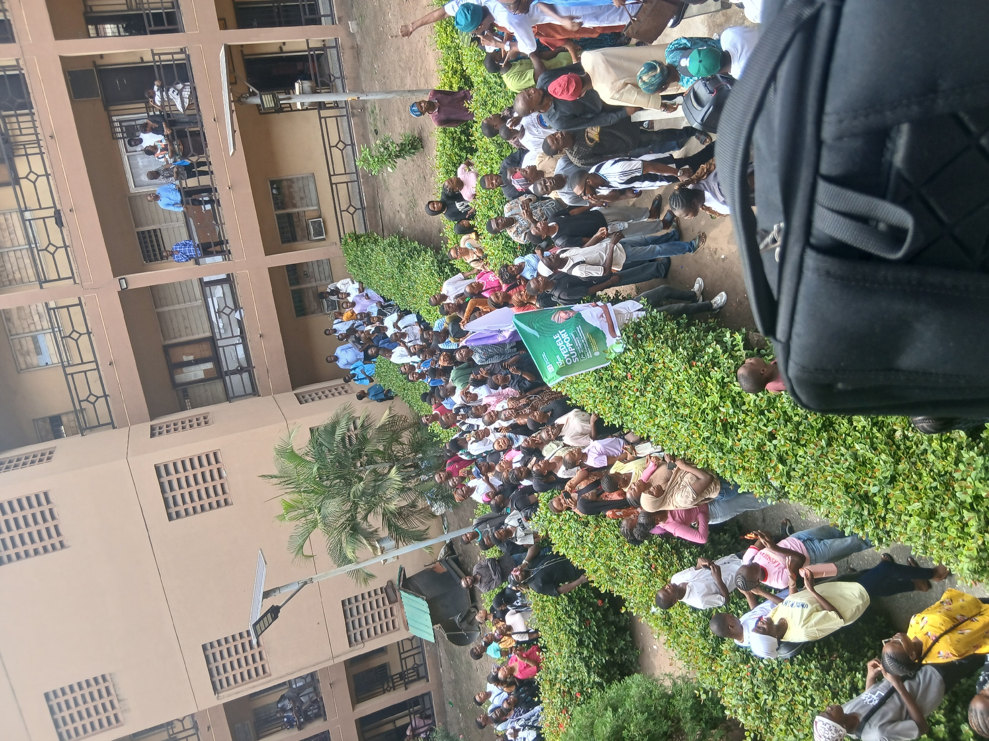 This image captures a large outdoor gathering of people, predominantly young adults, assembled in front of a multi-story building with a light brown facade. The setting, characterized by green hedges, a palm tree, and a street light with a solar panel, suggests an institutional or community environment, consistent with Lagos, Nigeria.
The crowd is densely packed, with individuals standing and some seated on the ground, wearing a variety of casual and some traditional attire. Several people are also visible observing from the balconies of the building. The general attention of the crowd appears to be directed towards a central point, implying an ongoing event such as a rally, lecture, or presentation.
A notable object is a green banner held within the crowd, featuring partially visible text that reads "OVERBLE SUPPORT" along with several logos below. The text likely forms part of a larger message related to an organization or cause.
The scene takes place during the daytime, under bright, clear weather conditions. A black backpack in the foreground suggests the perspective of someone situated within or very close to the gathering. The overall impression is one of an engaged and attentive crowd participating in a public event.