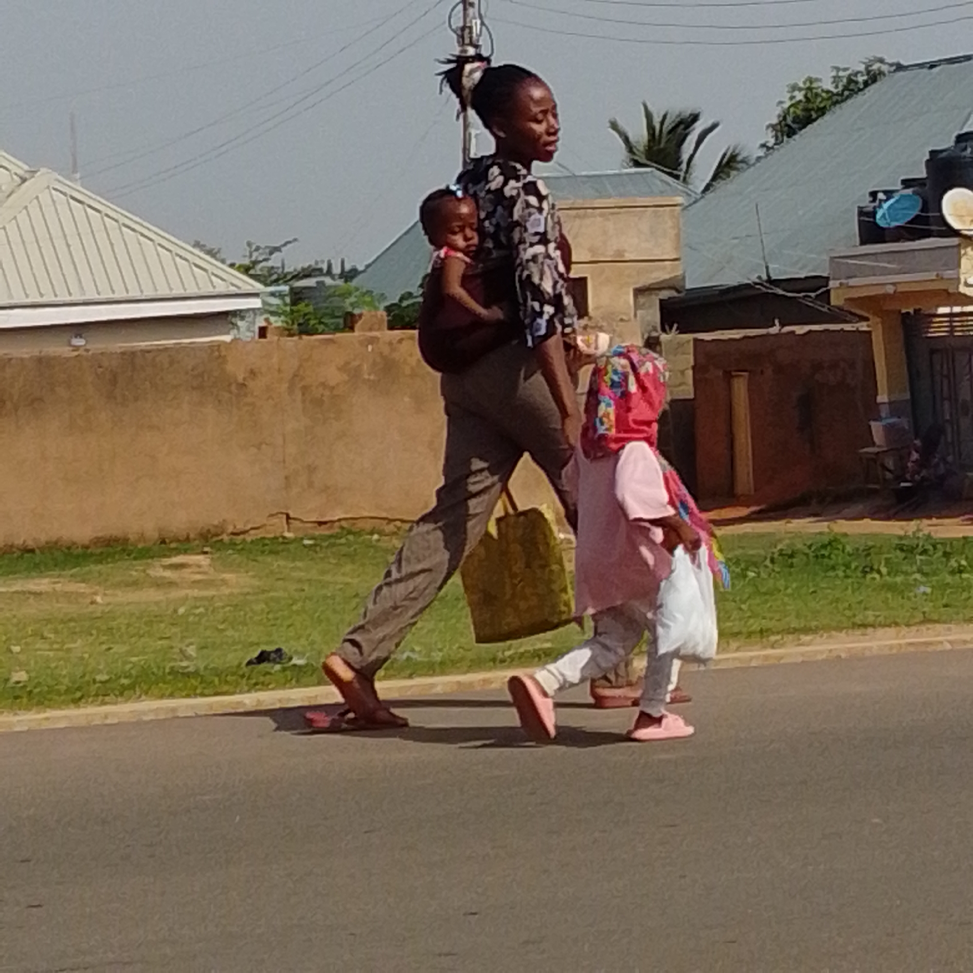 In Gombe, Nigeria, a woman walks on an asphalt road, carrying a baby on her back. She wears a patterned black and white top and grey trousers, with a large yellow tote bag slung over her shoulder. Beside her walks a young girl, also wearing a pink outfit and a patterned headscarf. The girl carries a white plastic bag. The background shows a dry brown wall, green grass, and buildings with corrugated metal roofs, consistent with urban architecture in the region. The sky is clear and bright, suggesting a sunny day.