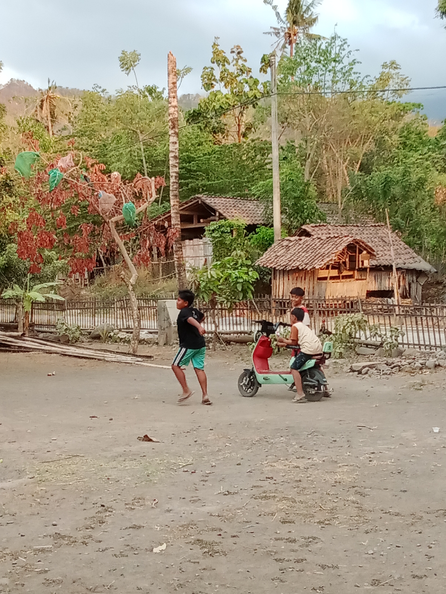 In Selowogo, Indonesia, a dusty, rural scene unfolds with three young boys. One boy in a black t-shirt and green shorts runs on the left side of the frame. To his right, two other boys are on a small, light-green and red scooter. One boy sits in front, wearing a light-colored sleeveless shirt and dark shorts, while the other boy sits behind him, wearing a white shirt and dark shorts. They are all positioned on a dirt ground. In the background, traditional Indonesian houses with thatched roofs are visible. The environment is characterized by lush green trees and vegetation, with hills in the distance under a cloudy sky. A wooden fence runs along the mid-ground, separating the houses from the open area. A lone tree with red and green items hanging from its branches stands to the left of the houses. The overall impression is of a simple, outdoor village setting.