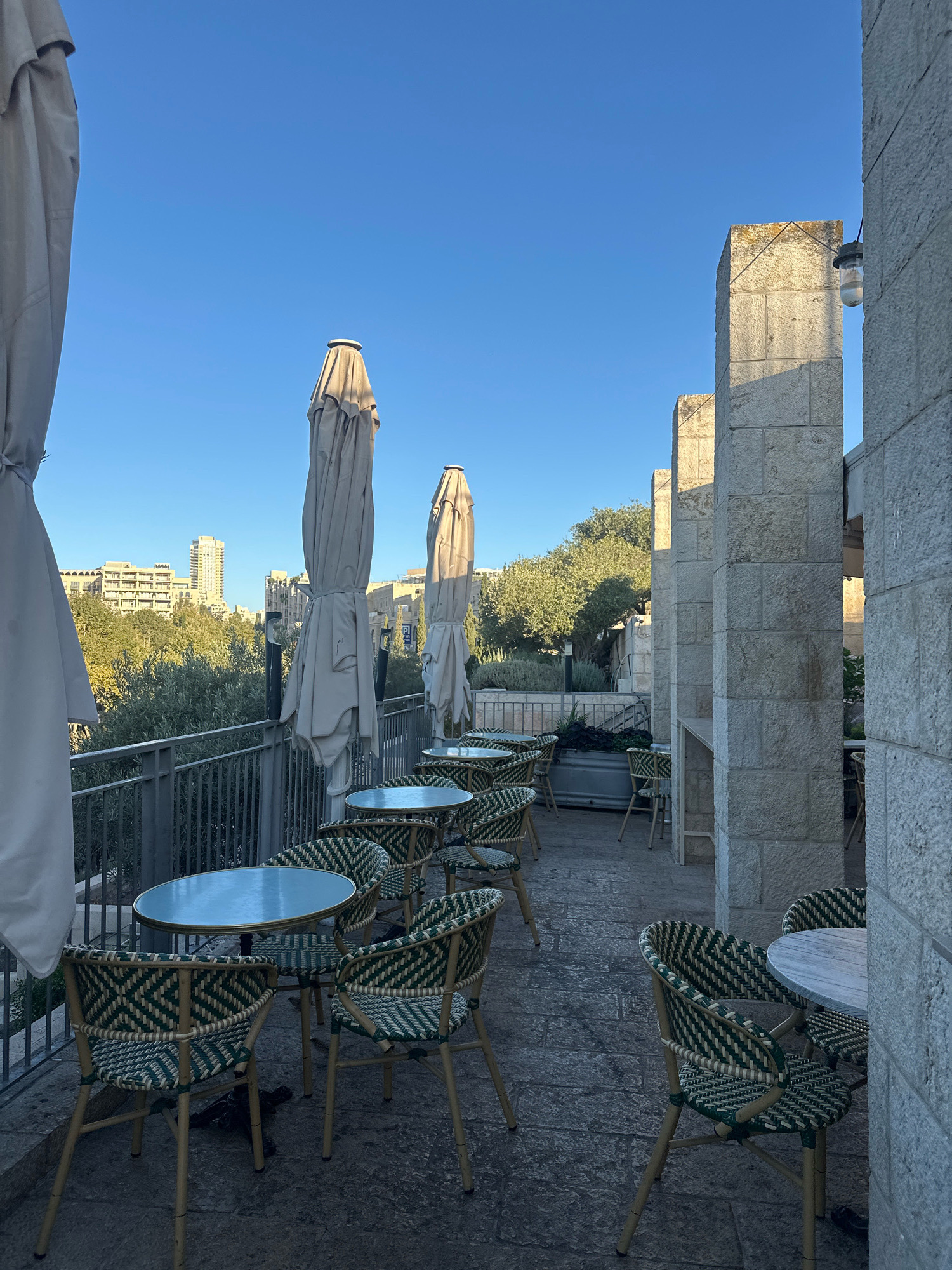 This image captures an outdoor seating area of a cafe or restaurant on a clear day in Jerusalem, Israel. The scene is dominated by a row of tables and chairs on a paved patio, overlooking a green landscape and city skyline in the distance. Several large, beige umbrellas are folded and tied, standing upright next to the tables. The chairs are woven with a distinctive green and white pattern, and the tables are round with light-colored tops.
To the right, a building constructed of rough-hewn, light-colored stone rises, with thick, square columns supporting an upper level or roof. A simple outdoor light fixture is visible attached to the stone wall. A metal railing separates the patio from a lower area with trees and shrubbery. In the background, several modern buildings, including a tall high-rise, are visible against a bright blue sky. The overall impression is one of a quiet, pleasant outdoor space, likely in a historic or upscale area of the city, given the architecture.