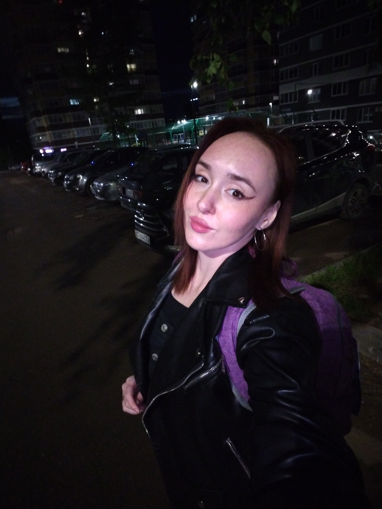 A young woman with dark red hair and fair skin poses for a selfie at night in Izhevsk, Russia. She is illuminated by an unseen light source, creating a glow on her face and highlighting her features. She has winged eyeliner and light pink lipstick, and is wearing hoop earrings. Her expression is calm with a slight smile. She is dressed in a black leather jacket over a dark top, with a purple backpack visible on her left shoulder.
The background shows an urban setting at night. Behind her, a row of dark-colored cars, including sedans and SUVs, are parked along what appears to be a street or parking lot. A multi-story building, likely an apartment complex, stands prominently in the background, with several windows lit, indicating residential or commercial activity. The overall scene is dark, characteristic of nighttime, with light sources primarily coming from the building windows and potentially streetlights off-camera, as well as the light illuminating the woman. A partially visible license plate on one of the cars reads "B826EK 177".