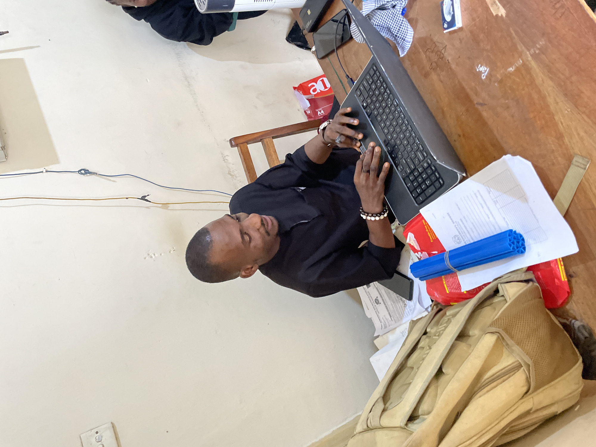 The image captures an indoor scene, likely an office or study area, in Durumi, Nigeria. A young man of African descent is seated at a wooden table, engrossed in using a laptop. He is wearing a black, long-sleeved traditional garment. His hands are on the keyboard and touchpad, suggesting he is actively working. He wears several bracelets on his left wrist, including one with white beads and another with darker beads, and a ring on his left index finger.
The table is cluttered with various items. To the left of the laptop is a beige backpack, partially obscuring a stack of papers. In front of the laptop are more documents, a blue cylindrical object bundled with tape, and a red package. To the right of the laptop, there are two smartphones, a black object that appears to be a power bank or external hard drive, and a neatly folded blue and white checkered cloth. A wooden chair is visible behind the man.
The background shows a plain, light-colored wall with some electrical fixtures and wires. The overall impression is of a functional but somewhat disorganized workspace. The lighting appears to be artificial.