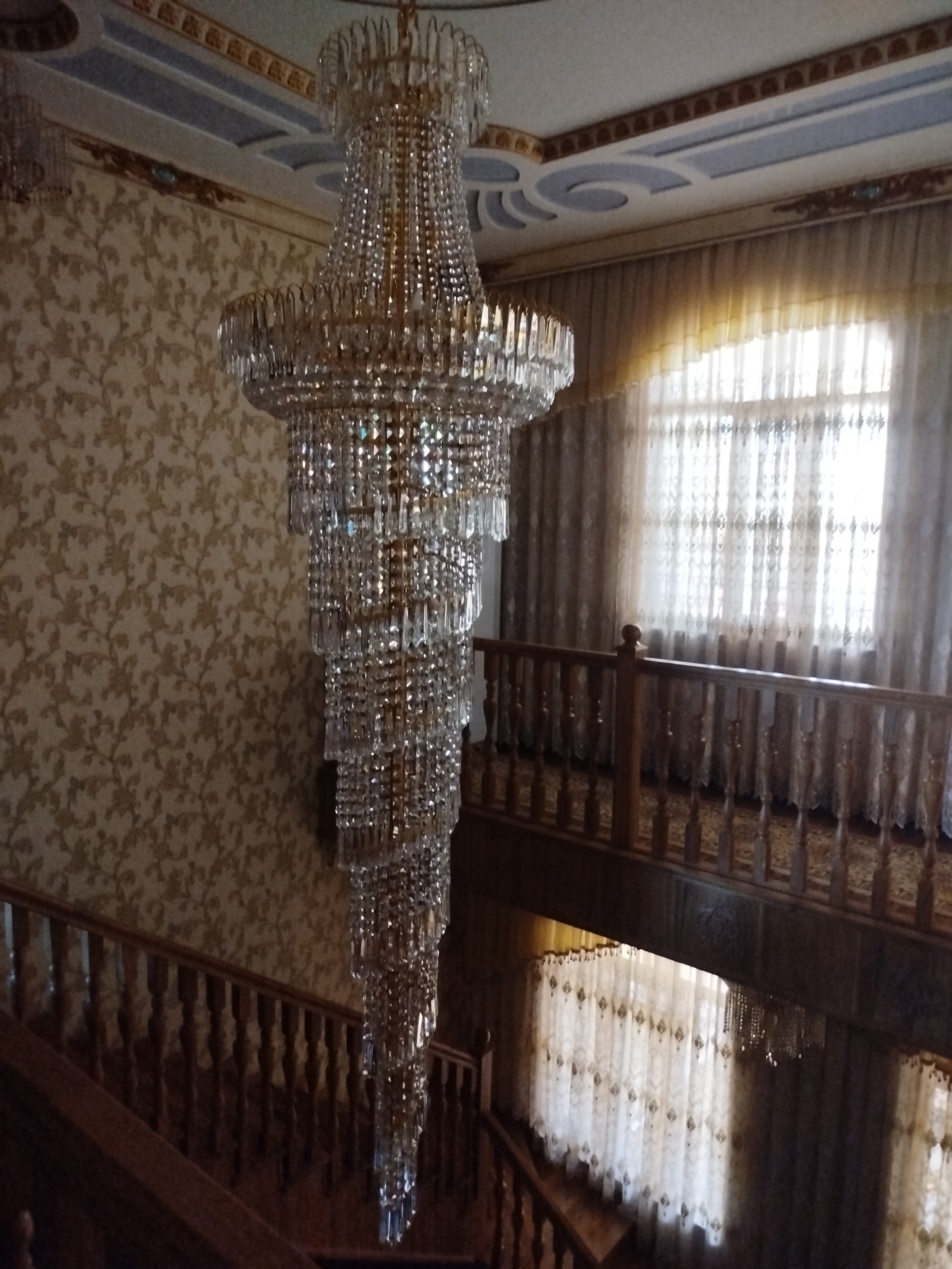 The image depicts a grand, multi-tiered crystal chandelier hanging in a richly decorated interior space, likely a hotel or historical building, in Bukhara, Uzbekistan. The chandelier is the central focus, with its numerous crystal pendants reflecting light.
The surrounding environment suggests opulence and traditional Uzbek design. To the left, a wall is covered with a textured, golden wallpaper featuring a subtle floral pattern. The ceiling exhibits ornate plasterwork with blue and gold accents, indicative of traditional Bukhara architectural styles.
In the background, sheer curtains with a delicate embroidered pattern cover a large window, allowing diffused natural light to enter the space. A wooden railing with intricately carved spindles runs along a staircase or balcony, framing the lower portion of the image. The overall impression is one of a luxurious and well-maintained interior, characteristic of the hospitality and historical significance of Bukhara. There are no people visible in the image.