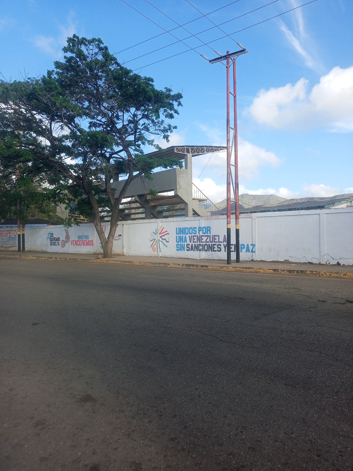This image depicts a street scene in Altagracia, Venezuela, featuring a large white wall adorned with political murals. On the left, a section of the wall displays a yellow banner with red text that reads "RESPETO PARA LA DIG..." followed by an illegible continuation. To its right, another mural shows text in Spanish, with an illustration of a hand making a peace sign and the words "LOS QUEREMOS DEVUELTA" and "NOSOTROS VENCEREMOS". Further to the right, the most prominent mural features large, bold Spanish text stating "UNIDOS POR UNA VENEZUELA SIN SANCIONES Y EN PAZ".
In the background, behind the wall, is a stadium-like structure with concrete bleachers and a metal framework. A tall, red utility pole with electrical insulators stands prominently on the right side of the frame, with power lines stretching across the blue sky. A large tree with dense green foliage partially obscures the stadium structure and the wall. The street in the foreground is asphalt, with a concrete curb and sidewalk running alongside the wall. The sky is mostly blue with scattered white clouds. The overall scene suggests a public space where political messaging is displayed.