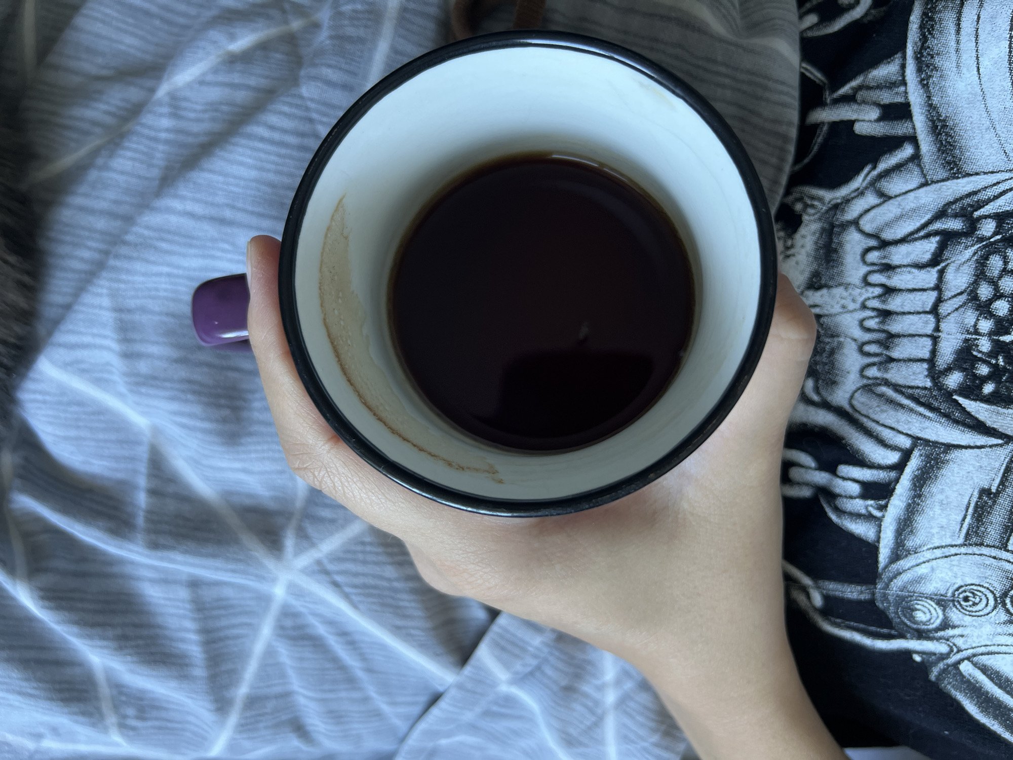 An overhead shot shows a hand holding a white mug with a dark liquid, possibly coffee, against a backdrop of gray, textured fabric. The mug has a black rim and a purple handle. A portion of a black t-shirt with a white skeleton print is visible at the bottom of the frame. The environment appears to be indoors, possibly on a bed or couch. There is no indication of the location being Tolyatti, Russia.